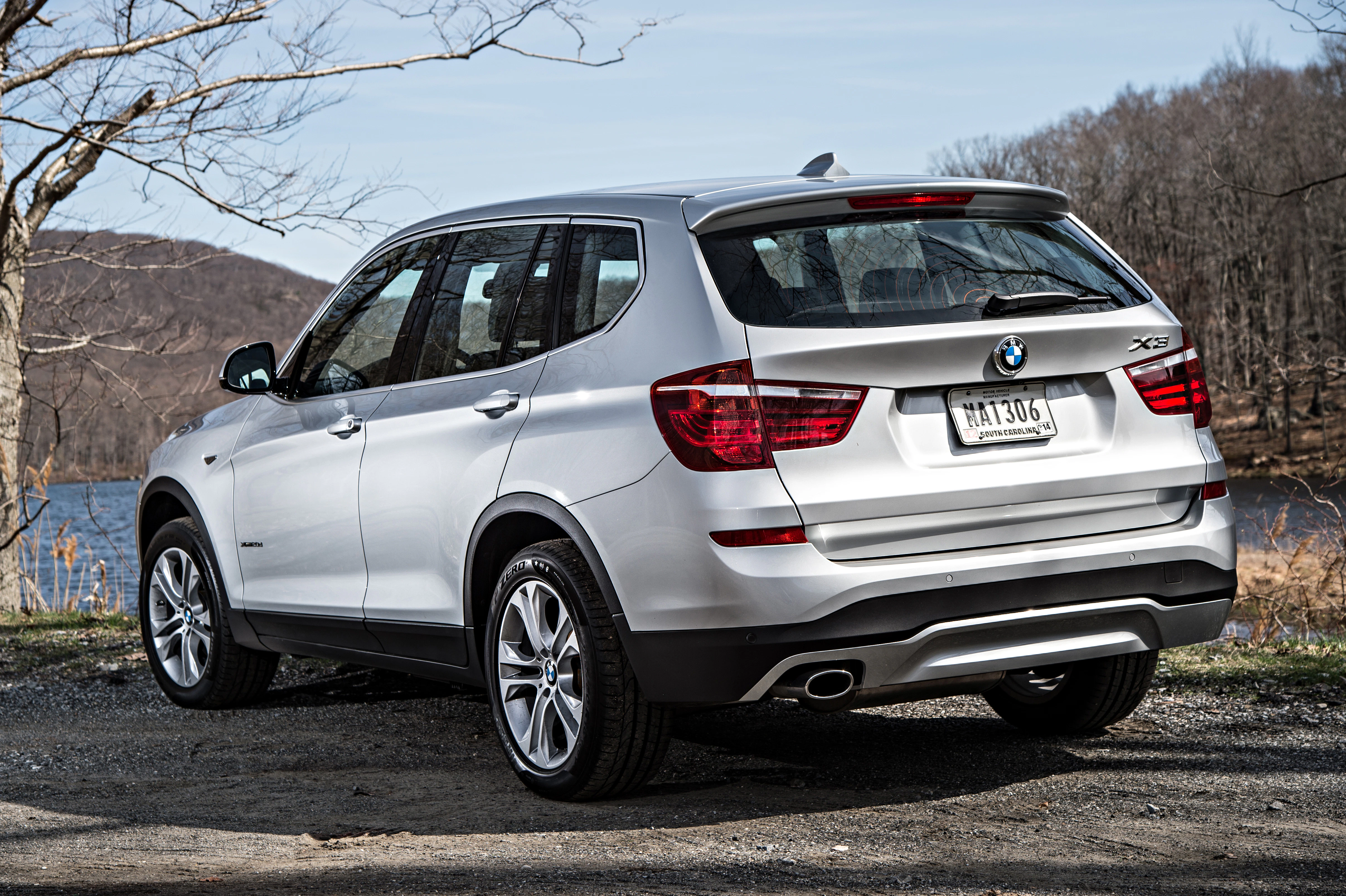 Silberner BMW X3 SUV steht auf einem Schotterweg nahe einem See, umgeben von kahlen Bäumen. Klarer Himmel im Hintergrund.