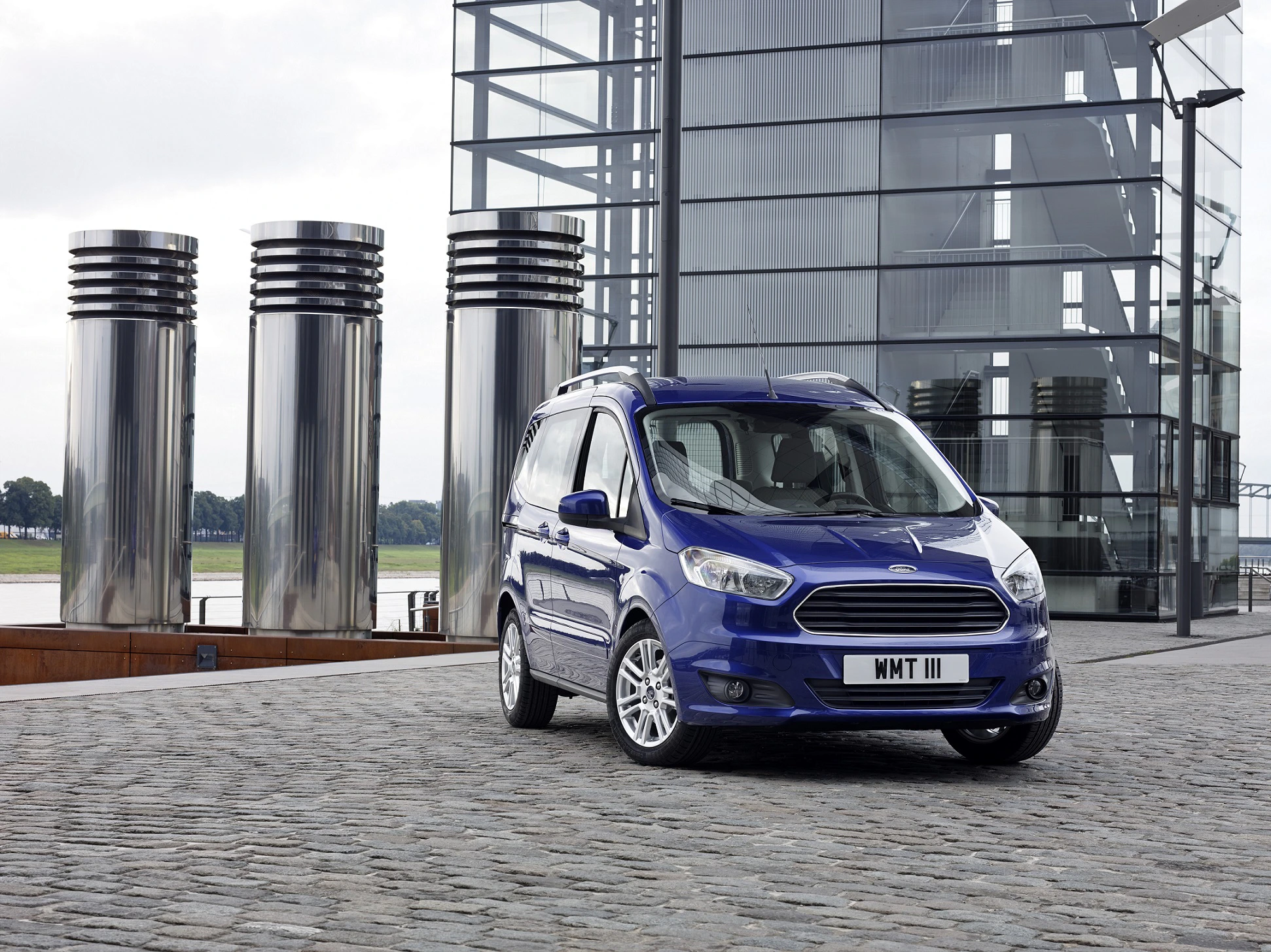Blauer Minivan parkt auf gepflastertem Platz vor modernem Glasgebäude mit silbernen Säulen im Hintergrund.