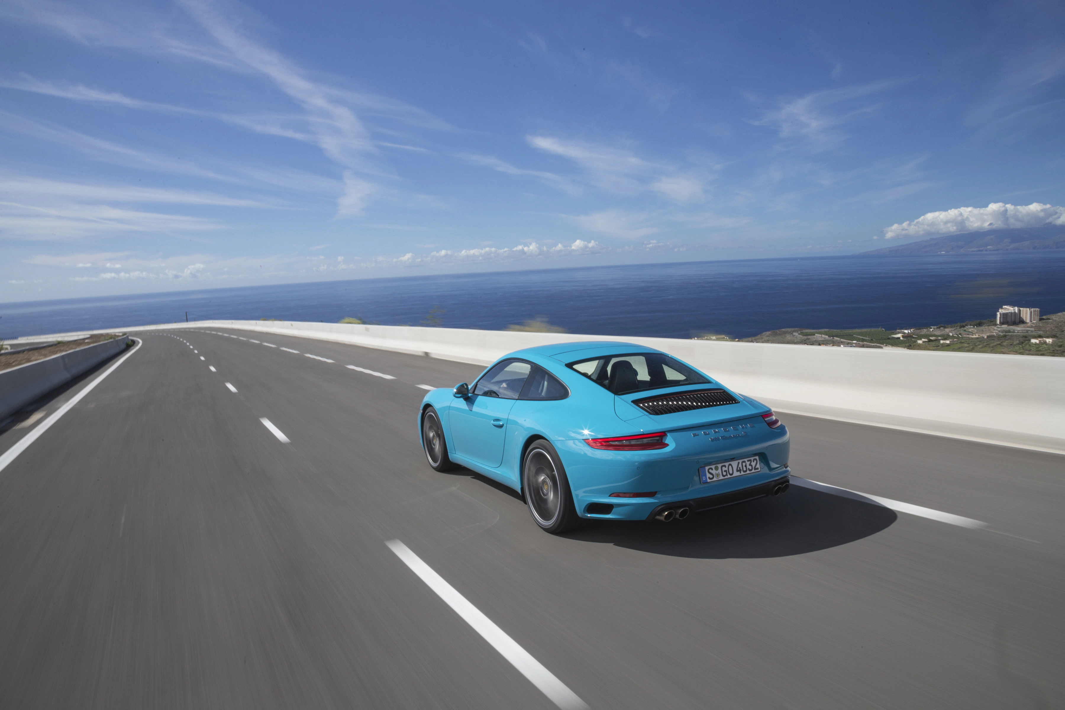 Blauer Sportwagen auf einer Küstenstraße; sonniges Wetter, Meer im Hintergrund, klare Himmel mit leichten Wolken.