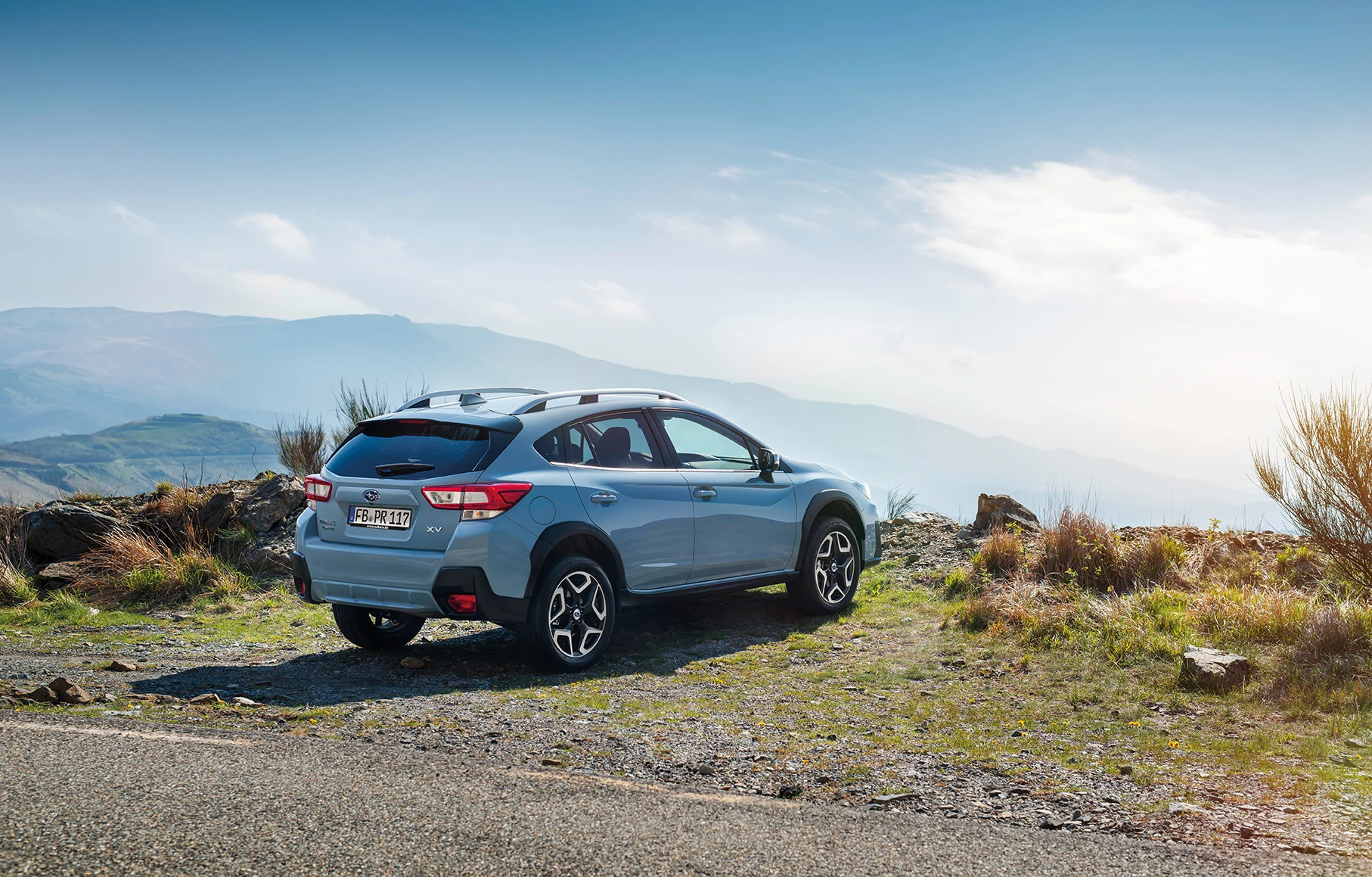Ein blauer SUV steht auf einer steinigen Anhöhe mit Aussicht auf ein bewaldetes Tal und einen klaren, wolkigen Himmel im Hintergrund.