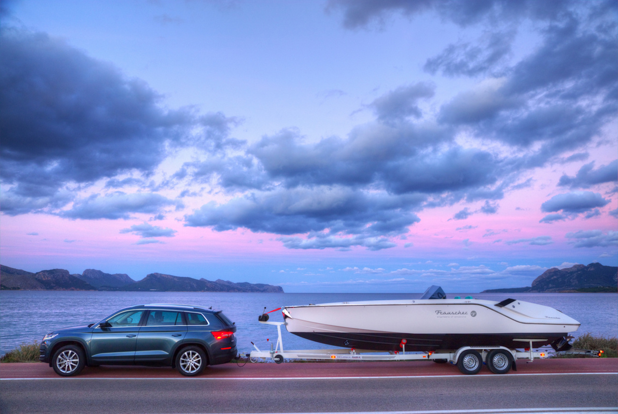 Auto mit Bootsanhänger auf einer Küstenstraße bei Sonnenuntergang, im Hintergrund Meer und bewölkter Himmel in Pastellfarben.