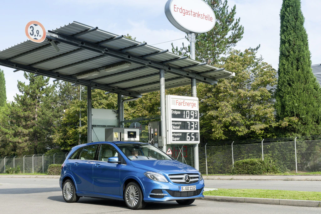Blaues Auto an einer Erdgas-Tankstelle mit Preisanzeige für Erdgas in Euro. Bäume im Hintergrund und eine Überdachung mit Solarpanel.