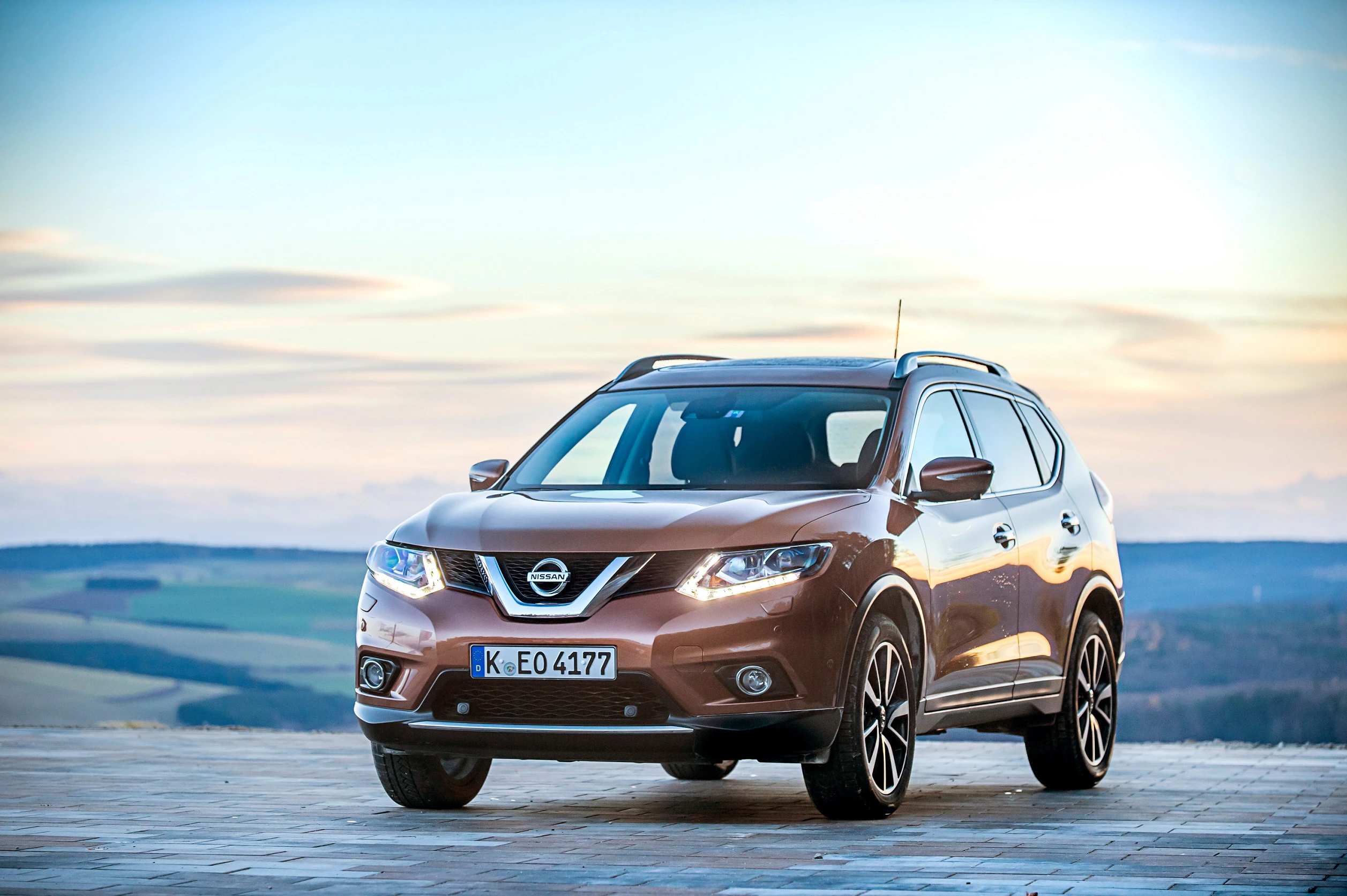 Brauner Nissan-SUV steht auf einer Steinfläche vor einer weiten Landschaft und Himmel bei Sonnenuntergang.