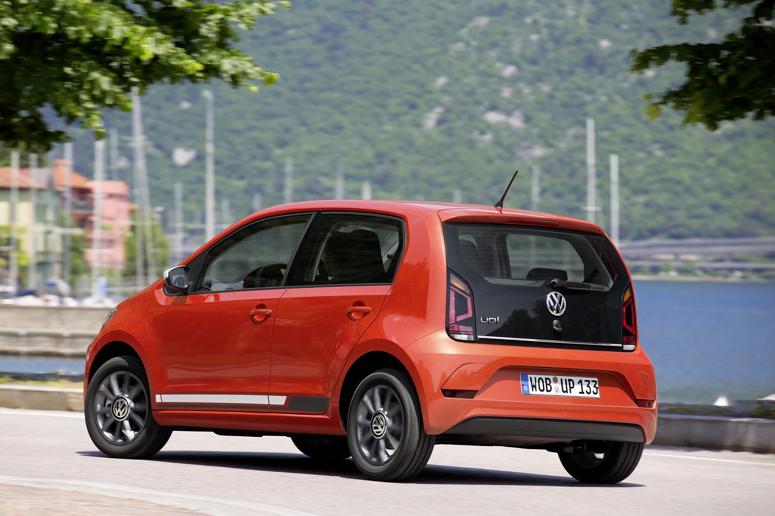 Roter Volkswagen Kleinwagen auf einer Straße nahe einem See, umgeben von grünen Bäumen und Hügeln im Hintergrund.