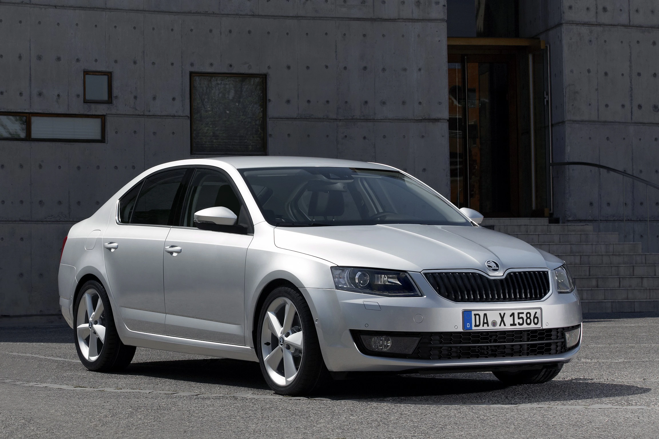 Silberner Škoda Octavia Limousine, geparkt vor einem modernen Gebäude mit Betonwänden und Treppe, an einem sonnigen Tag.
