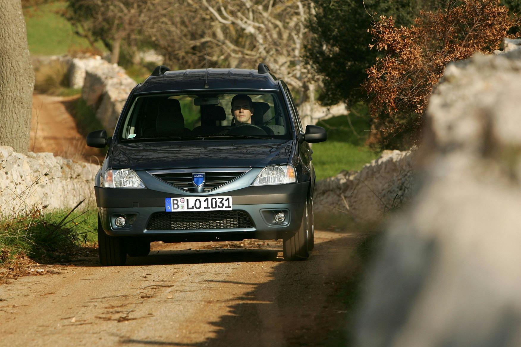 Auto fährt auf einem unbefestigten Weg zwischen Steinmauern. Fahrer trägt Sonnenbrille. Im Hintergrund Bäume und Gras.