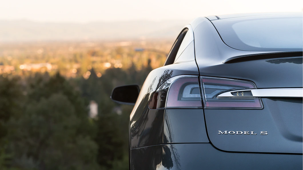 Nahaufnahme eines grauen Autos von hinten mit "Model S"-Emblem. Im Hintergrund ist eine unscharfe Landschaft zu sehen.