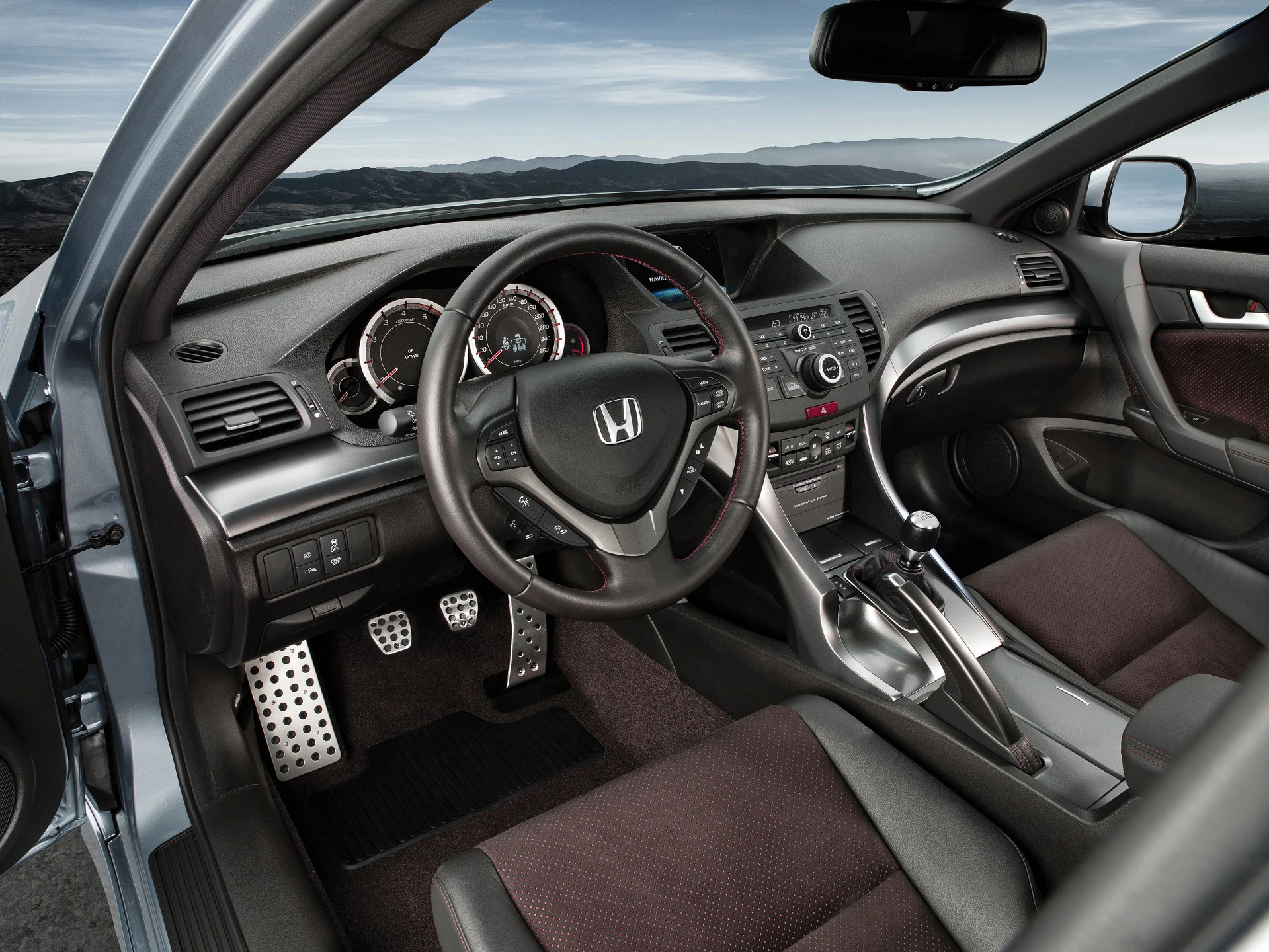 Innenraum eines modernen Honda-Autos mit schwarzem Lenkrad, Armaturenbrett, Schalthebel und digitalem Tacho, umgeben von grauen Sitzen.