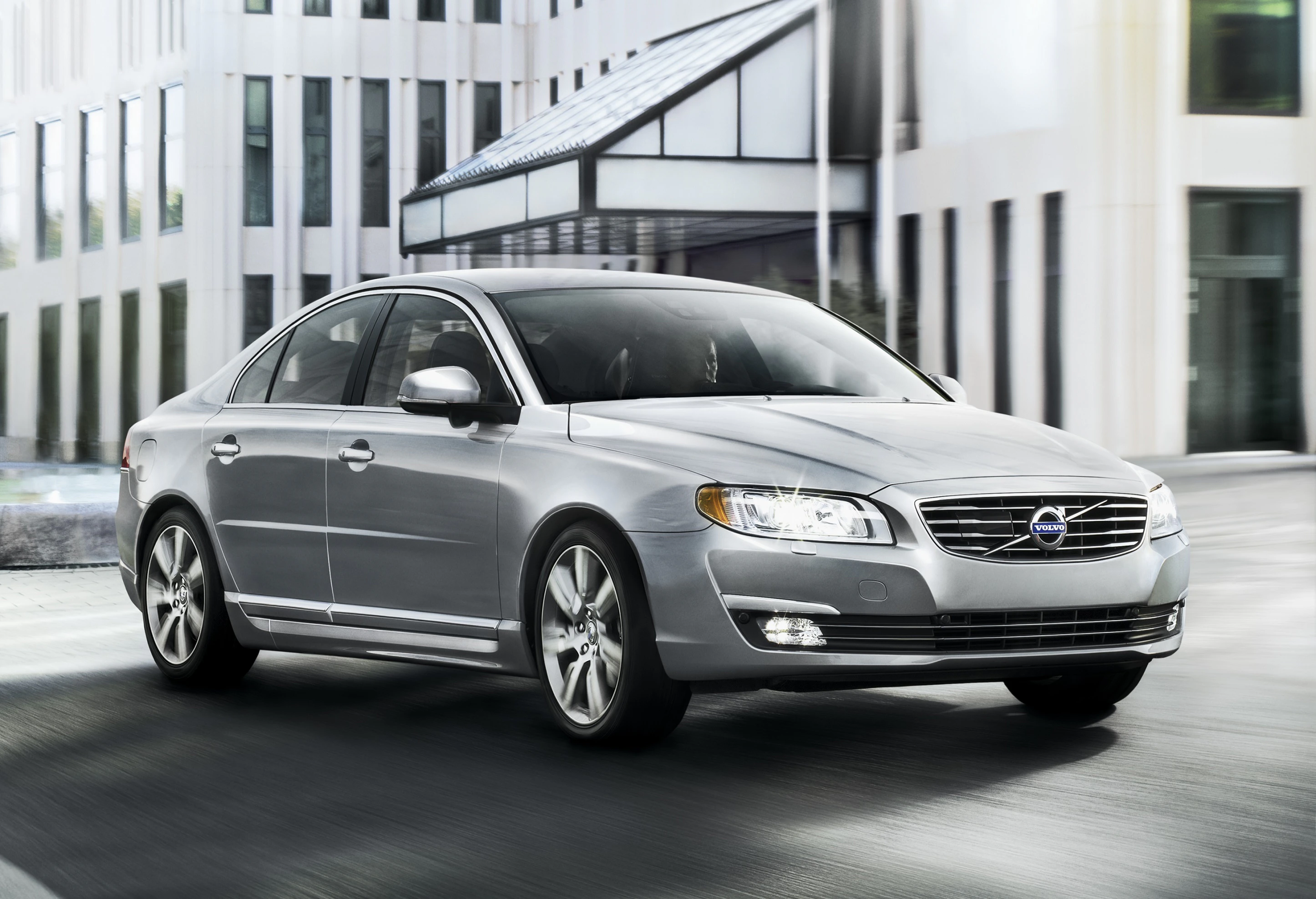 Silberner Volvo S80 fährt in urbanem Umfeld, moderne Architektur im Hintergrund, klare Linien und elegantes Design des Fahrzeugs.