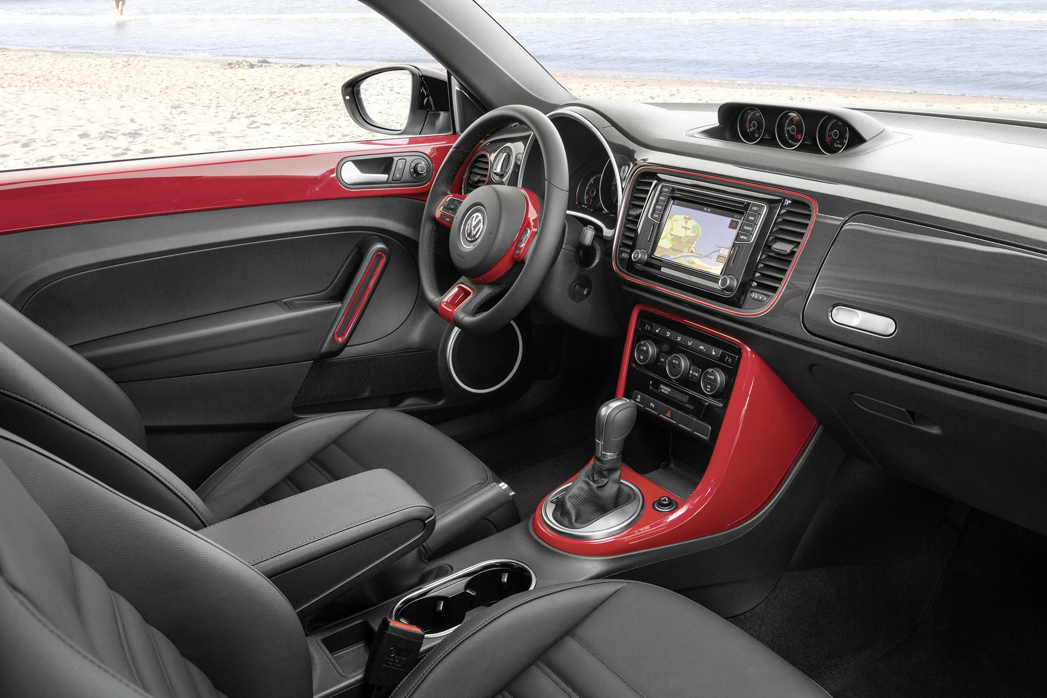 Innenraum eines Autos mit rotem Armaturenbrett. Sicht auf Lenkrad, Schalthebel und Navigationssystem. Fenster zeigt Strand im Hintergrund.