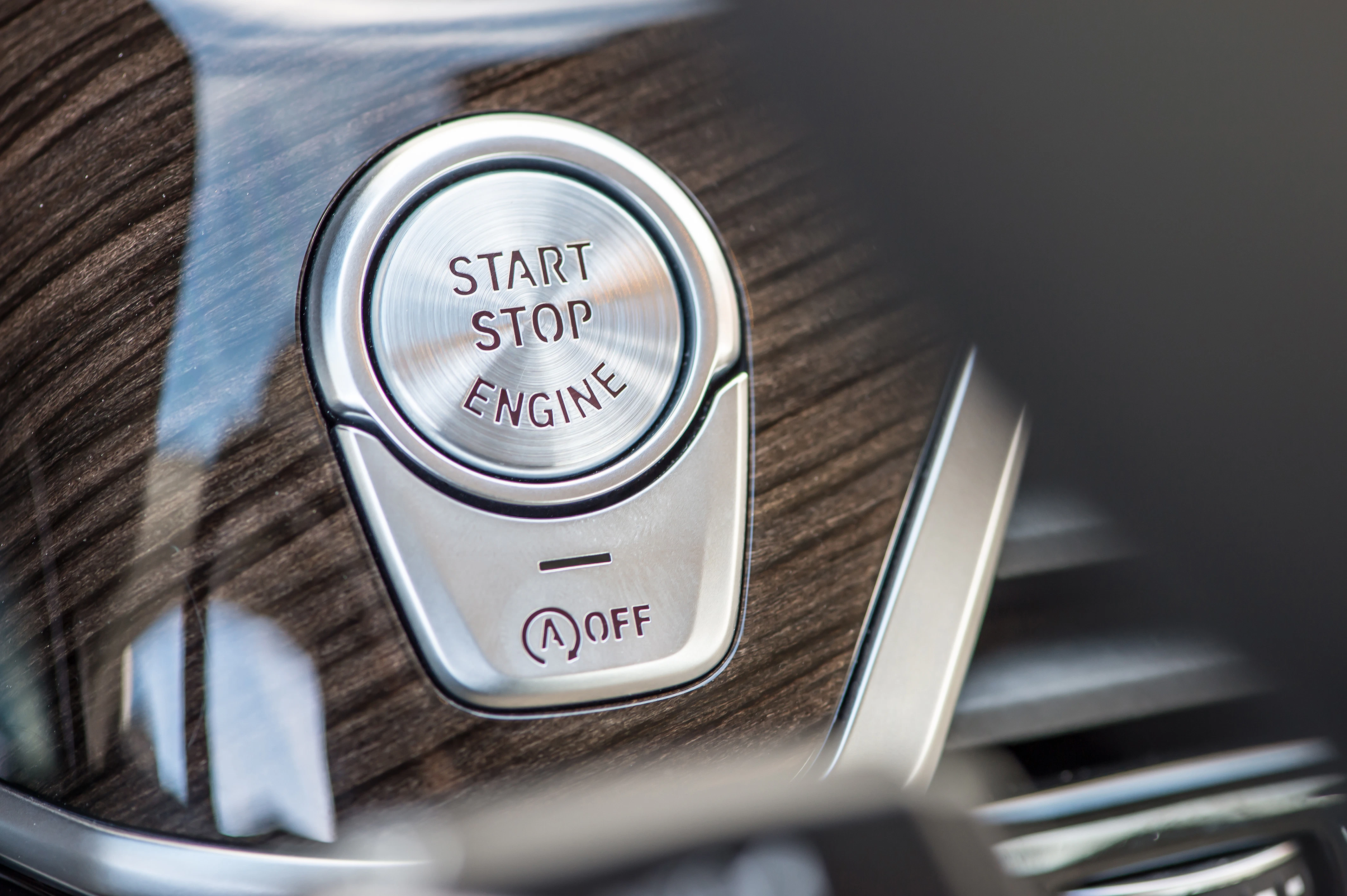 Nahaufnahme eines silbernen Start-Stopp-Motorknopfs in einem Auto, umgeben von hölzerner Armaturenbrettverkleidung.