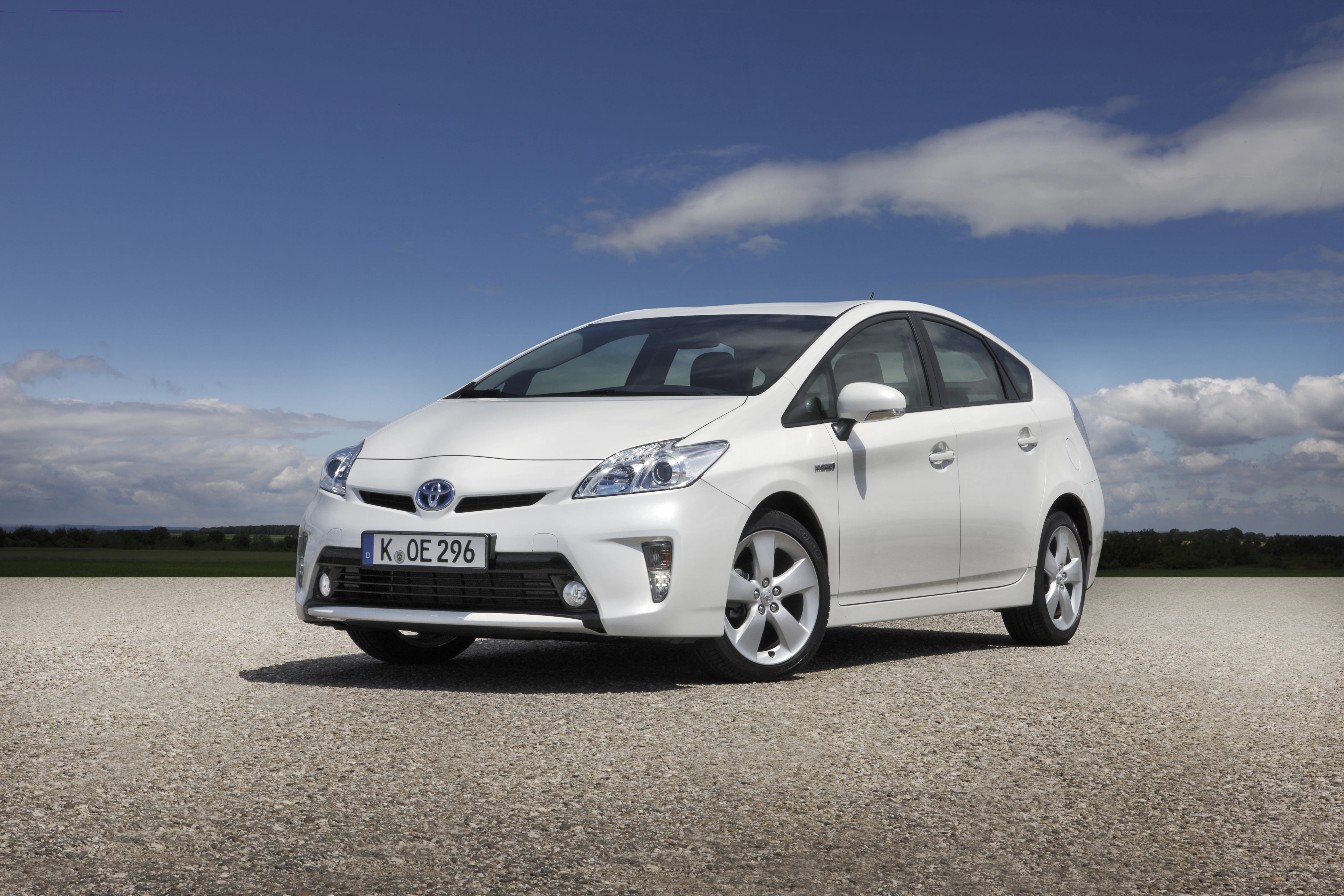 Weißer Toyota Prius steht auf einer Straße vor blauem Himmel und Wolken. Lizenznummer K OE 296 sichtbar.