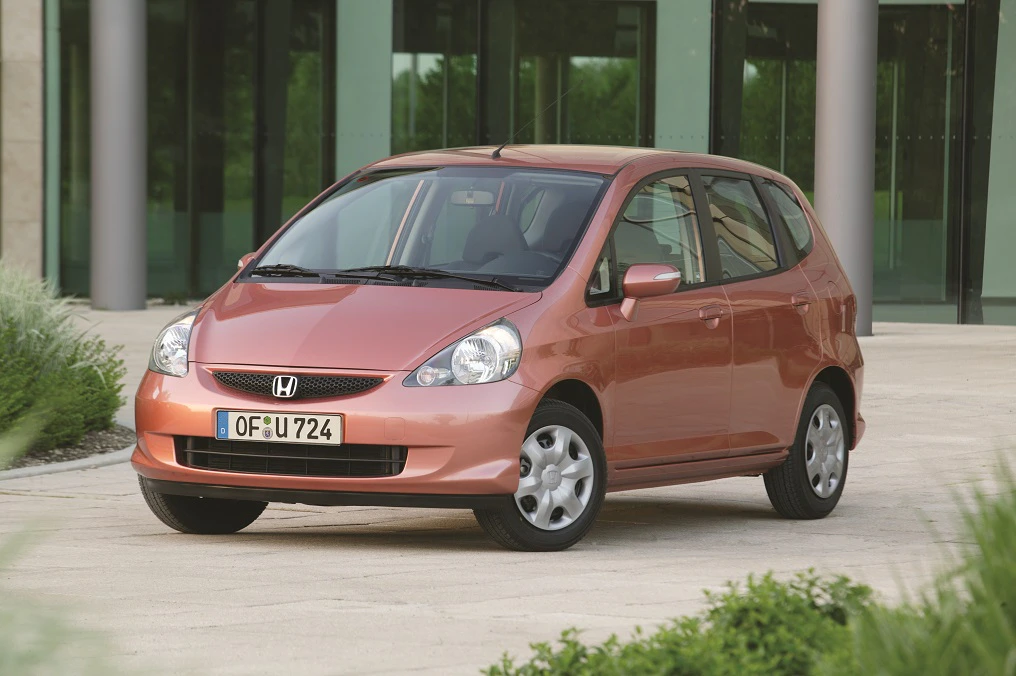 Ein orangefarbener Honda Jazz steht vor einem modernen Gebäude mit Glastüren, umgeben von grünen Pflanzen.