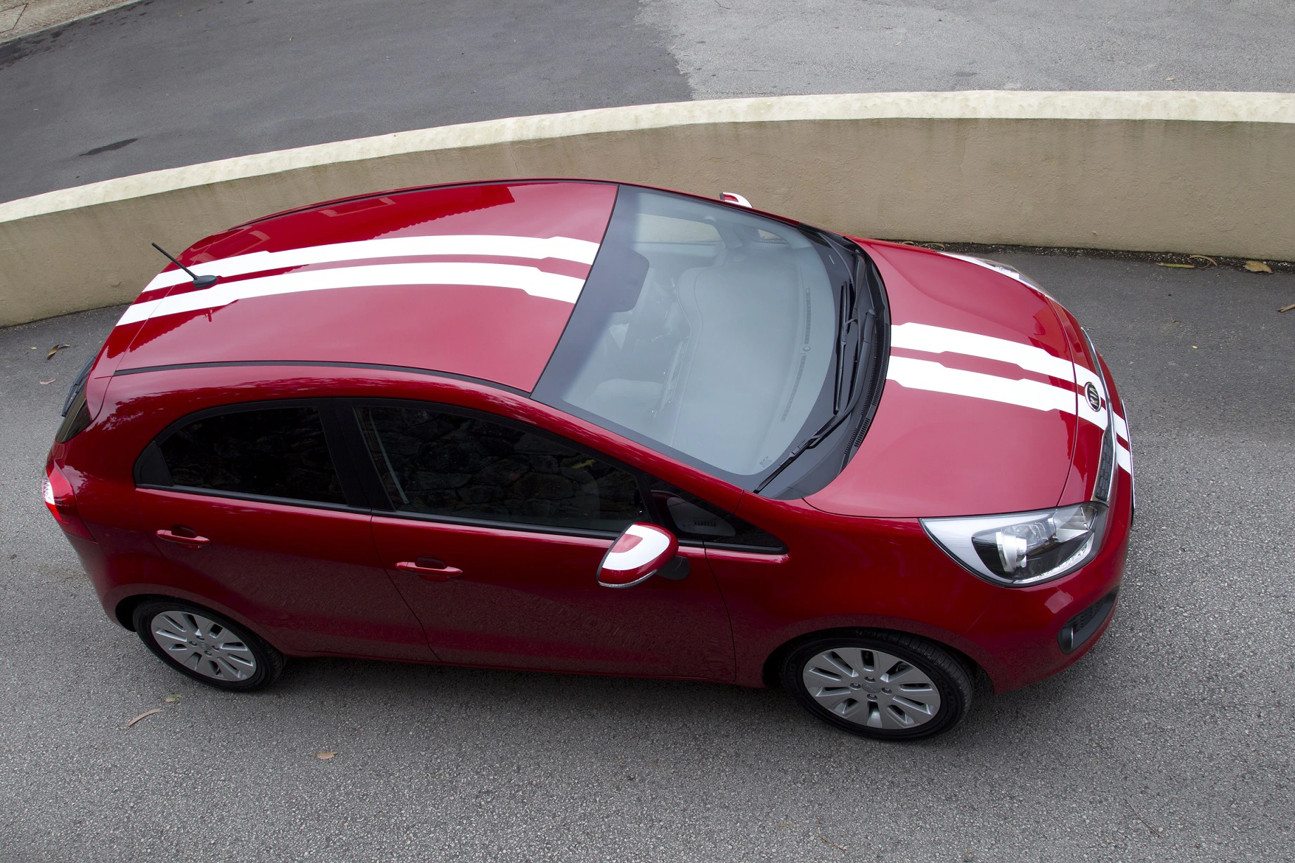 Rotes Auto mit weißen Streifen, seitliche Draufsicht. Das Fahrzeug steht auf grauem Asphalt neben einer beige gemauerten Mauer.