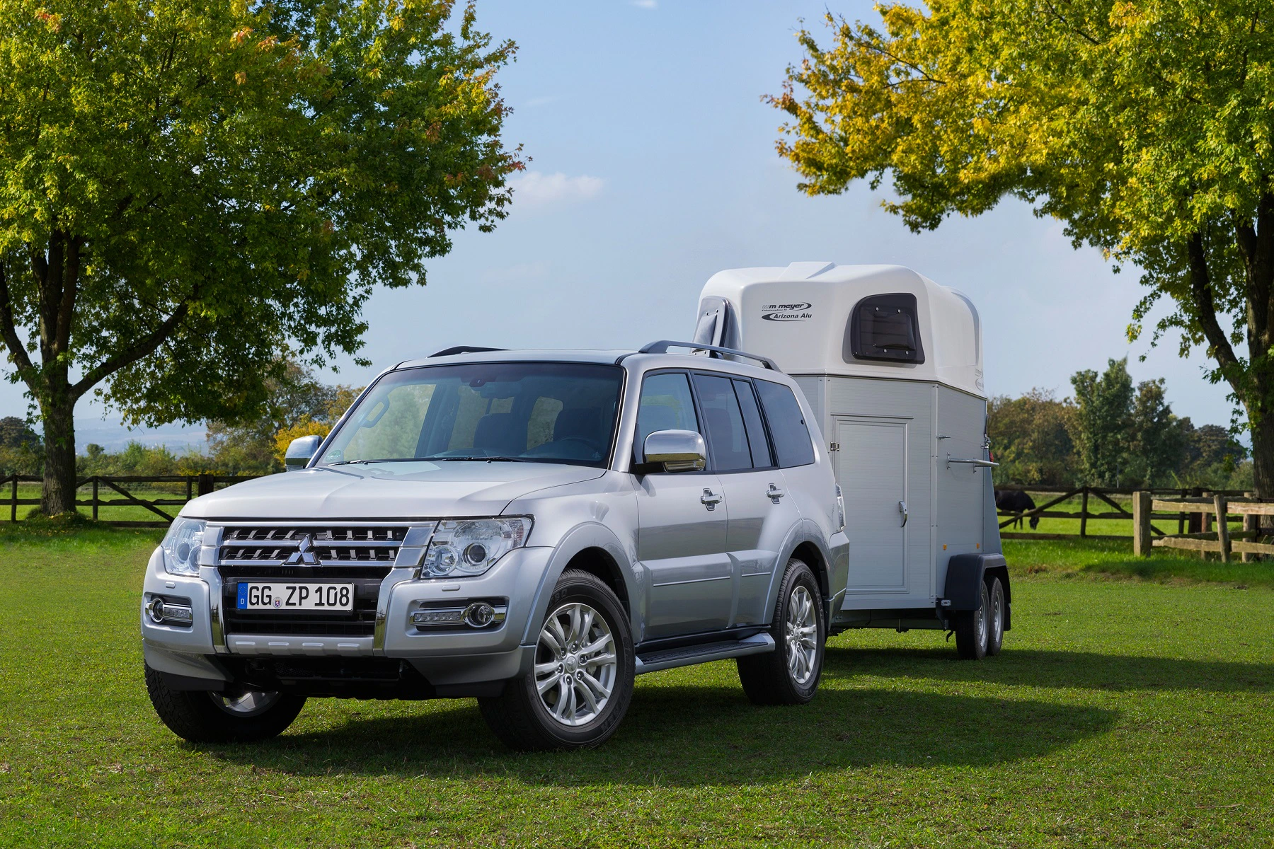 Silberner SUV mit Anhänger steht auf einer grünen Wiese, umgeben von Bäumen und Holzzaun im Hintergrund, unter einem klaren Himmel.