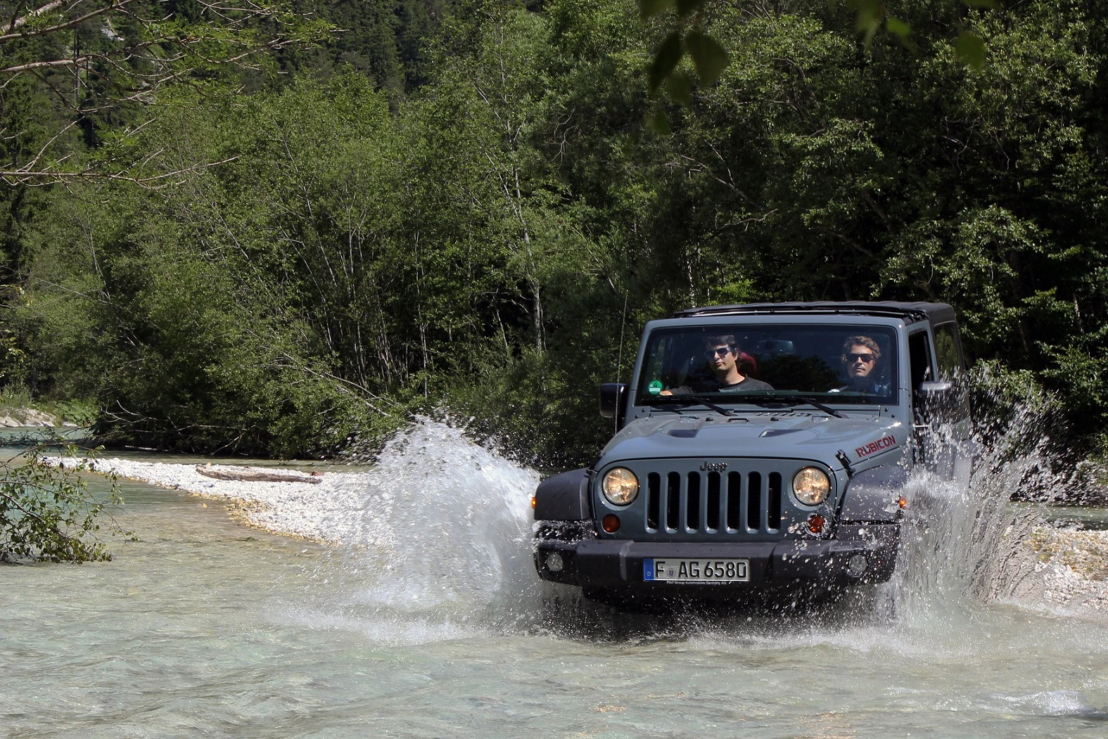 Jeep fährt durch einen flachen Fluss, spritzt Wasser auf. Zwei Personen sitzen im Wagen. Wald im Hintergrund.