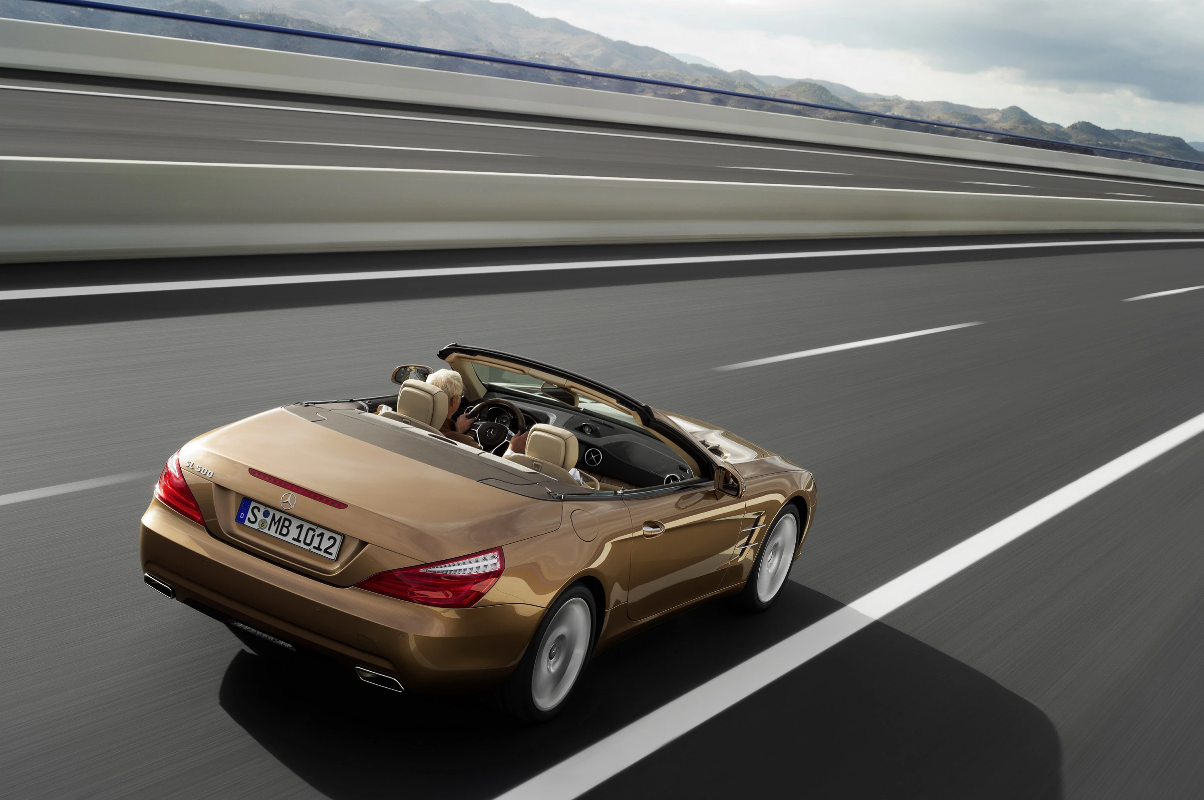 Goldenes Cabriolet fährt auf einer Autobahn. Offen und ohne Personen bei sonnigem Wetter, Berge im Hintergrund. Sportliches Design.