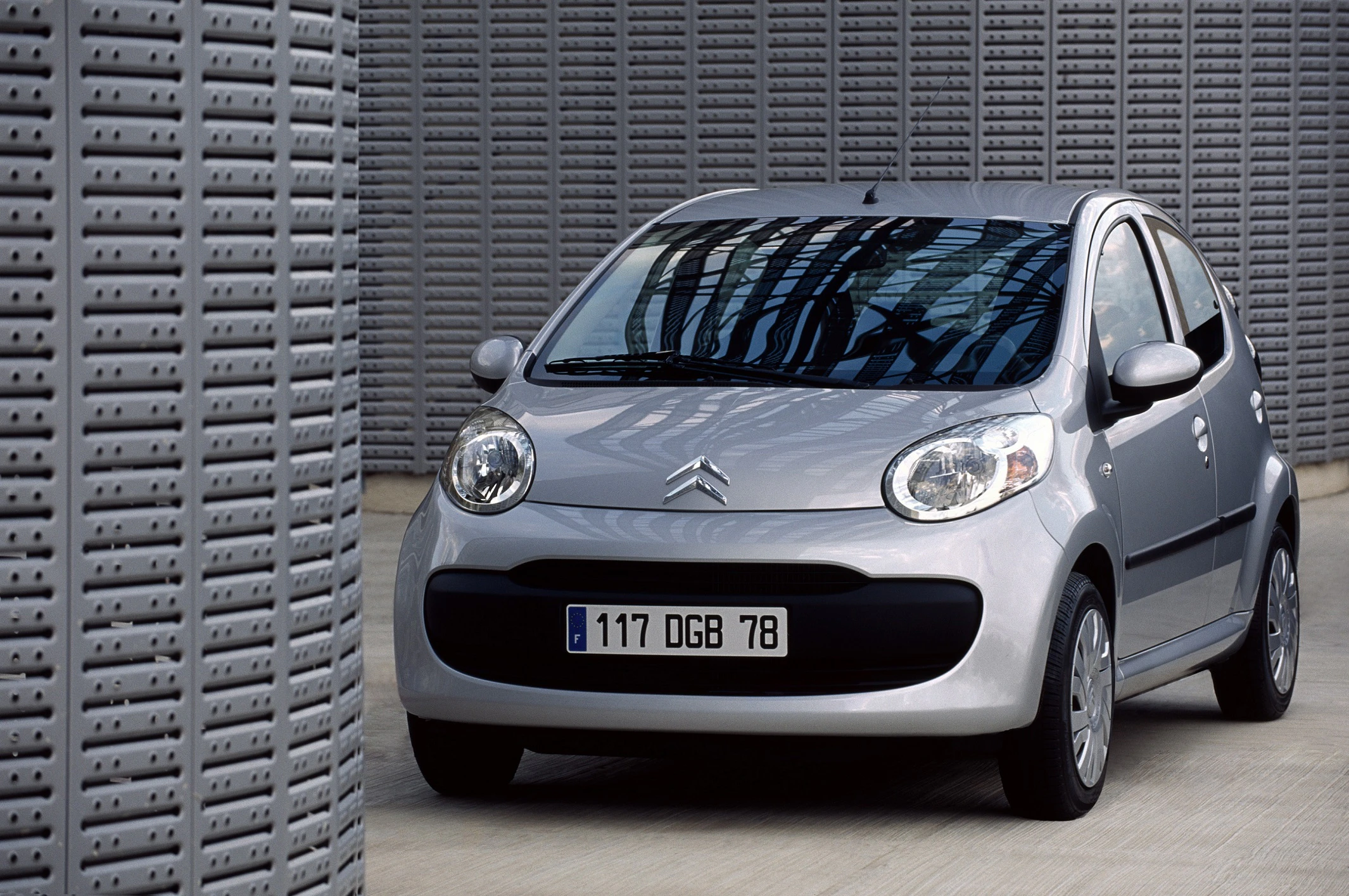 Silberner Citroën Kleinwagen vor einer modernen, grauen Wand mit geometrischen Mustern. Fahrzeug ist frontal zu sehen.
