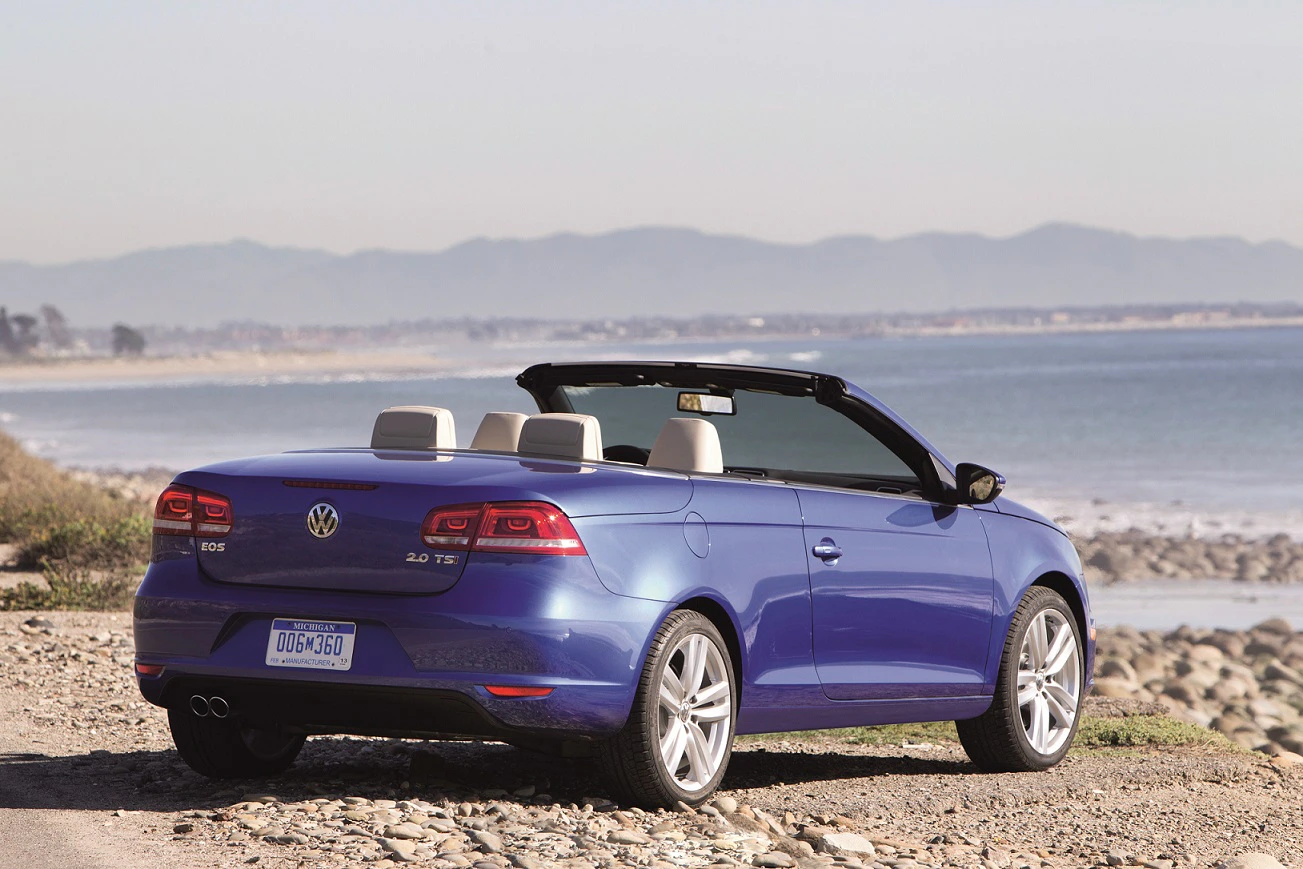 Blaues VW-Cabrio parkt an einem steinigen Strand mit Blick aufs Meer, Berge im Hintergrund, sonniges Wetter, Verdeck geöffnet.