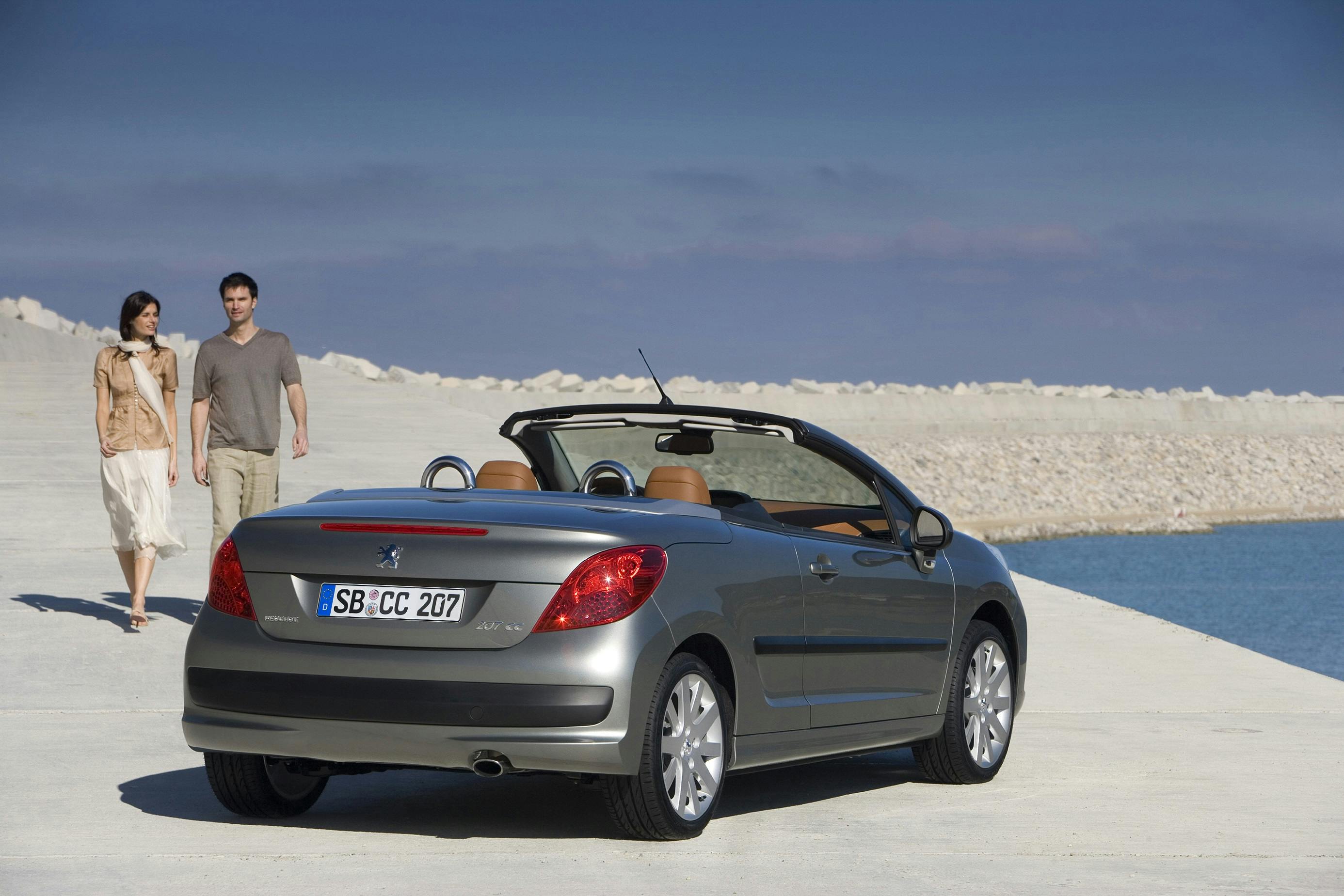 Ein silbernes Cabriolet steht auf einem sonnigen Pier mit Blick aufs Meer. Ein Paar geht in legerer Kleidung am Ufer entlang.