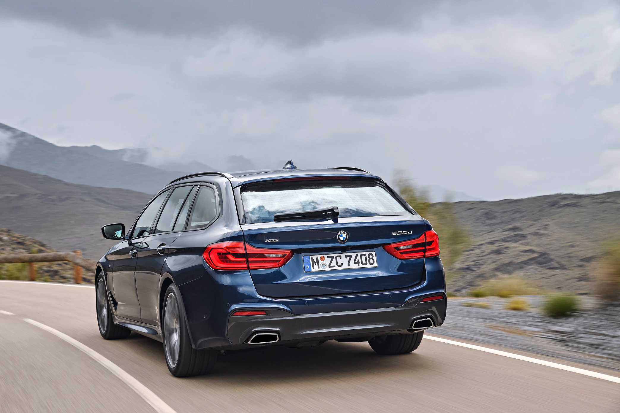 Ein blauer BMW Touring fährt auf einer Landstraße, umgeben von bergiger Landschaft unter bewölktem Himmel.