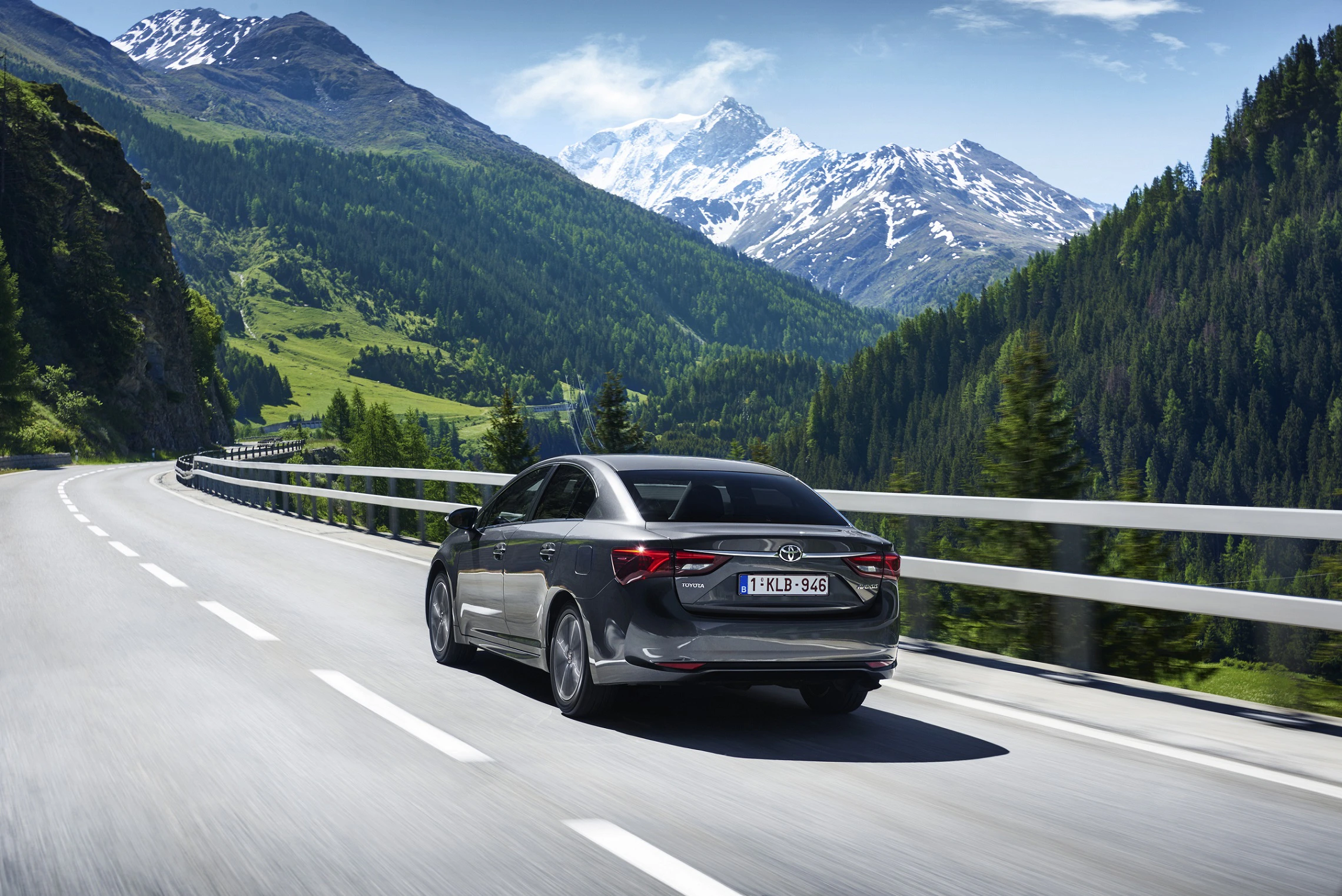 Ein schwarzes Auto fährt auf einer kurvigen Straße durch eine malerische Berglandschaft mit schneebedeckten Gipfeln und grünen Wälder.