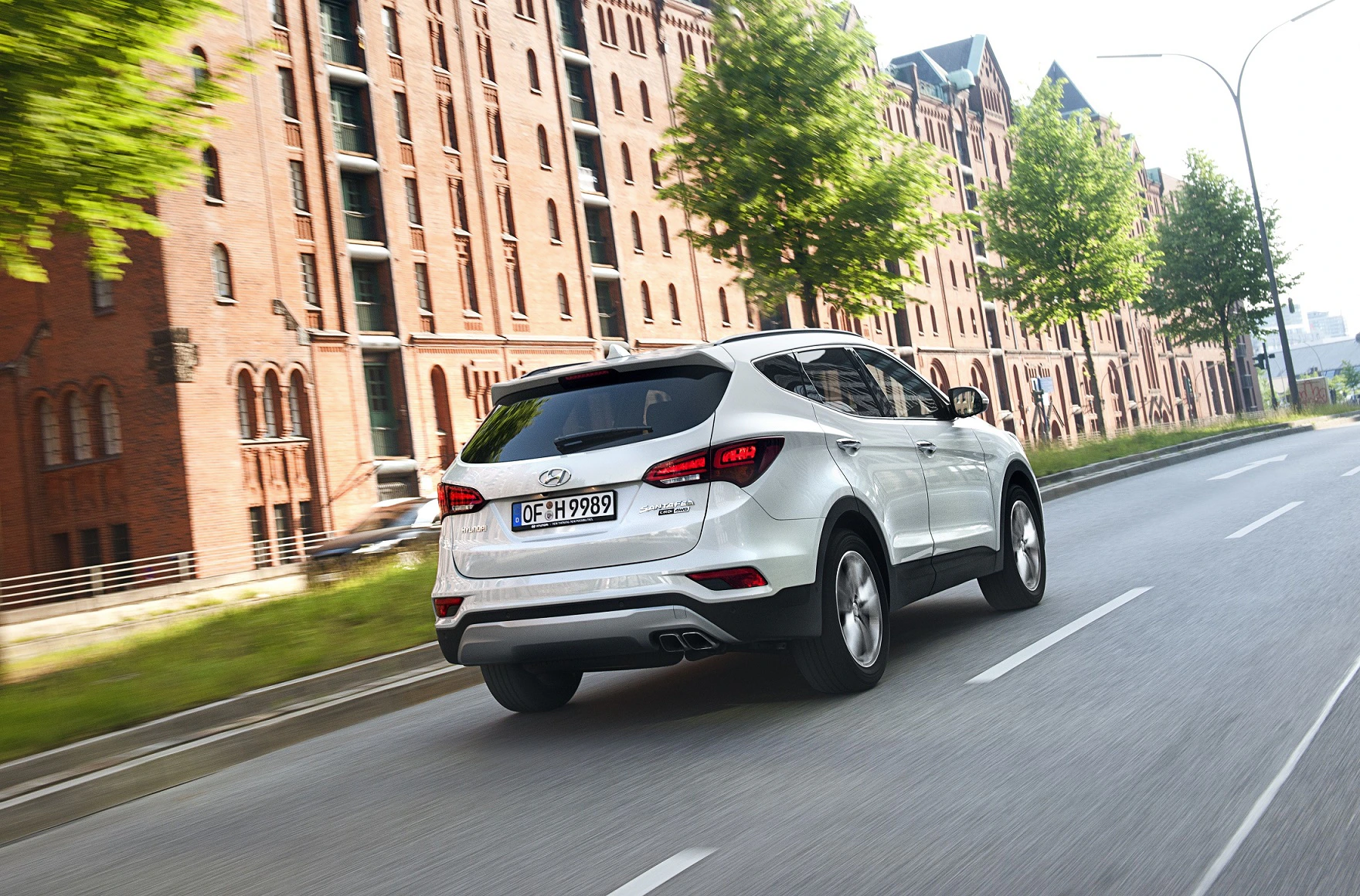 Ein weißes SUV fährt auf einer städtischen Straße mit Backsteingebäuden und grünen Bäumen im Hintergrund.