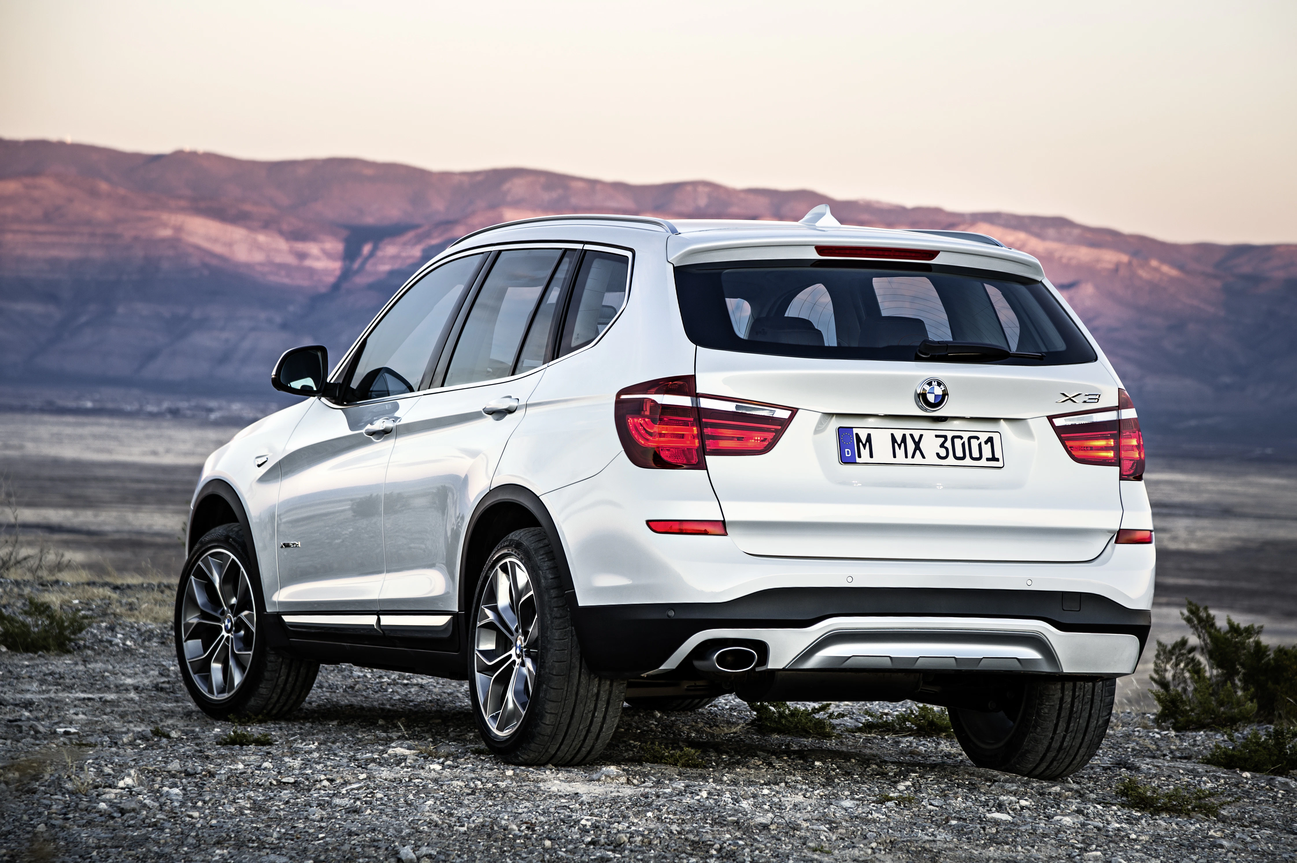 Weißer BMW SUV steht in einer kargen Landschaft vor einer Gebirgskulisse bei Sonnenuntergang.