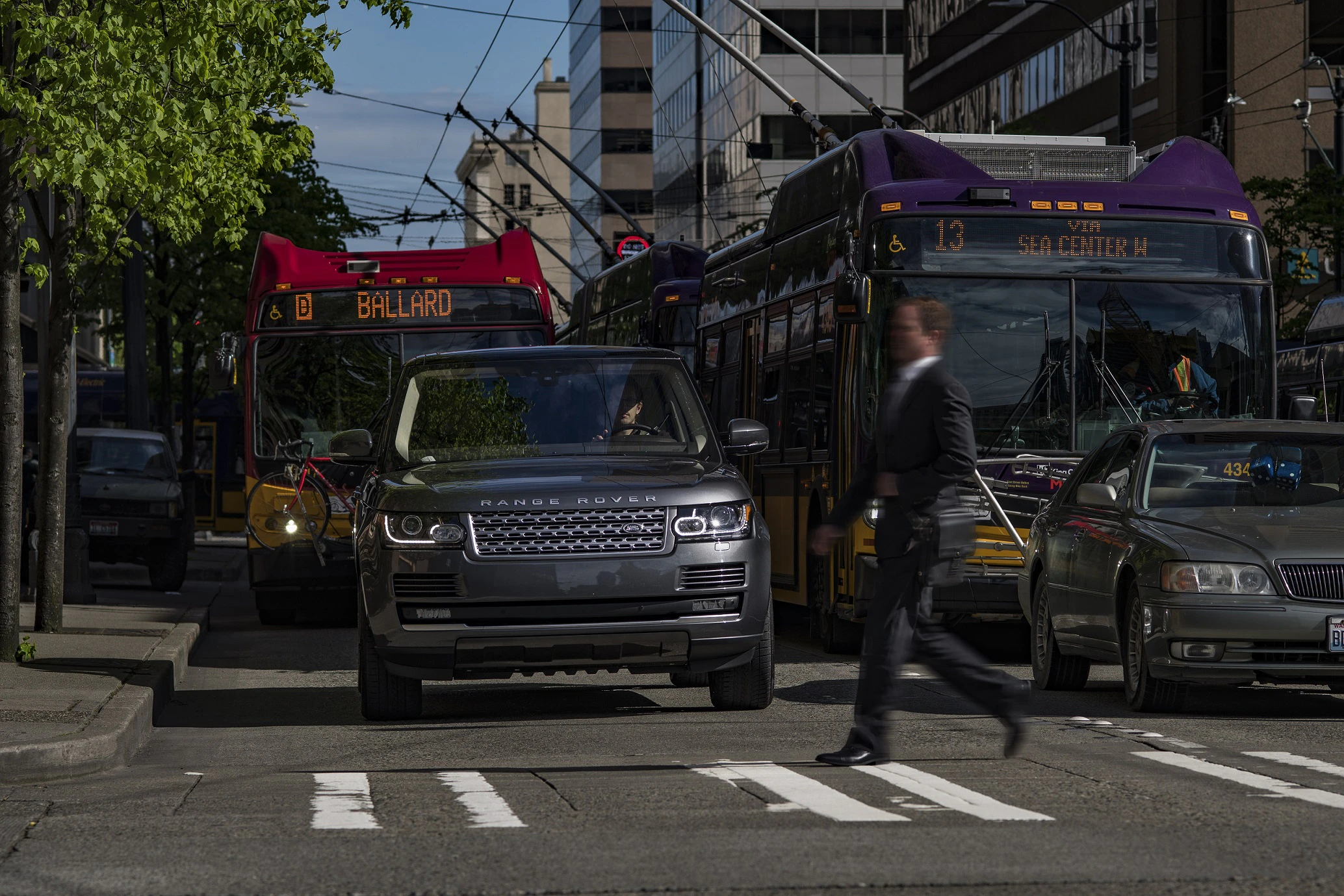 Ein Mann in Anzug überquert eine belebte Straße vor einem grauen Range Rover und Bussen in einer städtischen Umgebung.