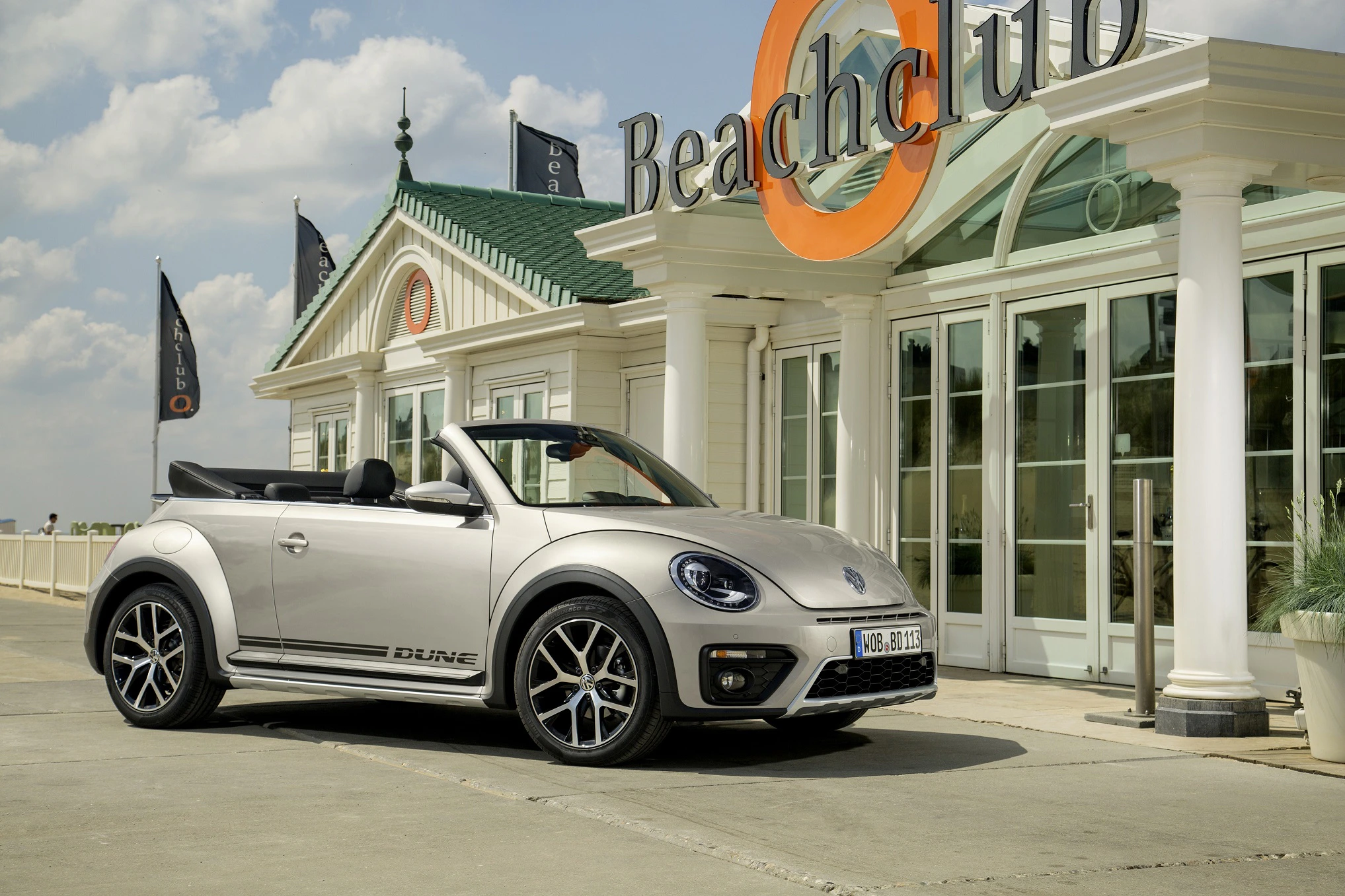 Silbernes VW Beetle Cabrio parkt vor einem Beachclub-Gebäude bei sonnigem Wetter. Retro-Design mit modernen Elementen.