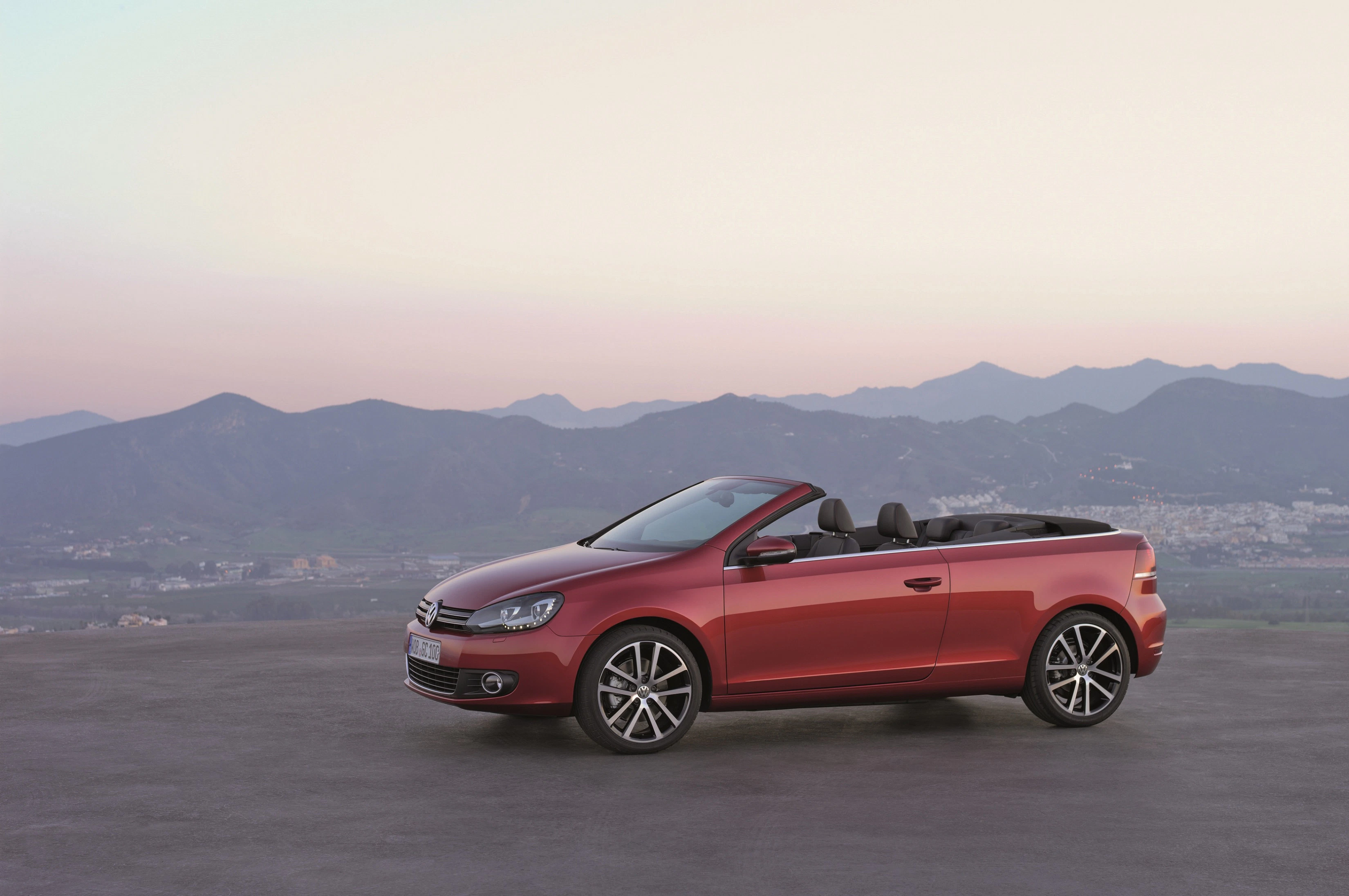 Rotes Cabrio vor bergiger Landschaft bei Sonnenuntergang. Himmel in Pastelltönen, Auto auf Asphaltfläche geparkt.