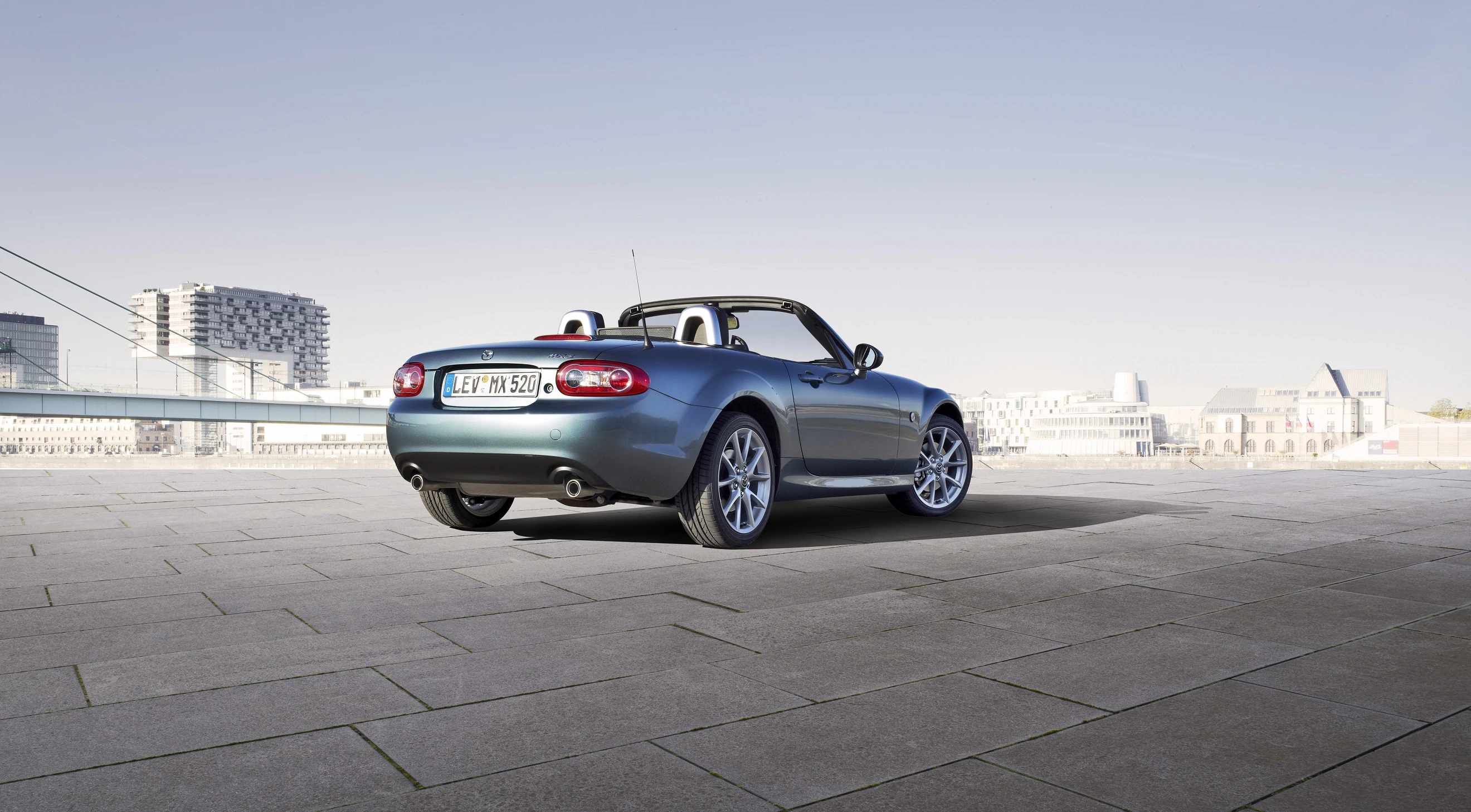 Blauer Cabrio-Sportwagen steht auf einem gepflasterten Platz, im Hintergrund moderne Gebäude und blauer Himmel.