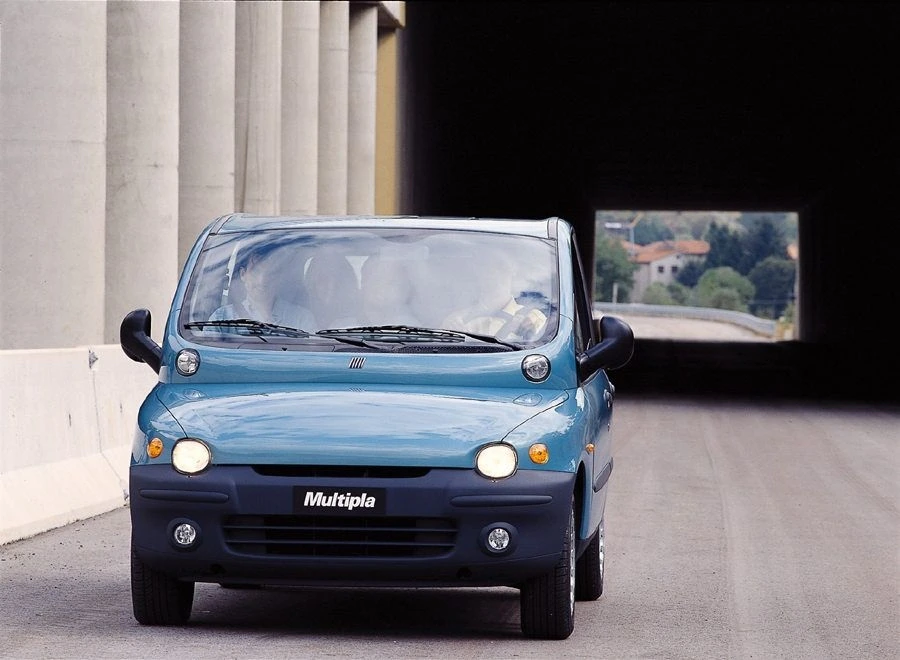 Blauer Fiat Multipla fährt auf einer Straße aus einem Tunnel. Im Hintergrund sind Bäume und Häuser zu sehen.