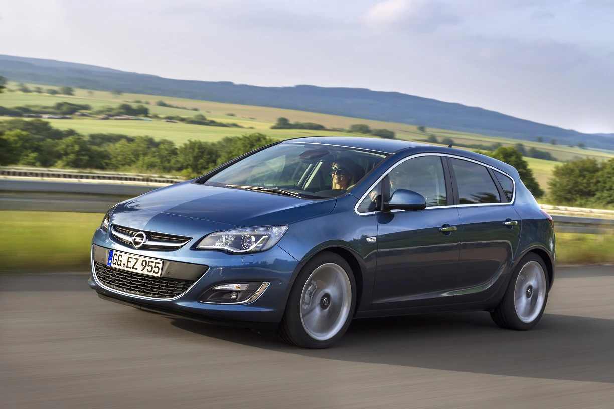 Blauer Opel Astra fährt auf einer Landstraße, umgeben von grünen Feldern und Hügeln im Hintergrund. Klarer Himmel mit einigen Wolken.