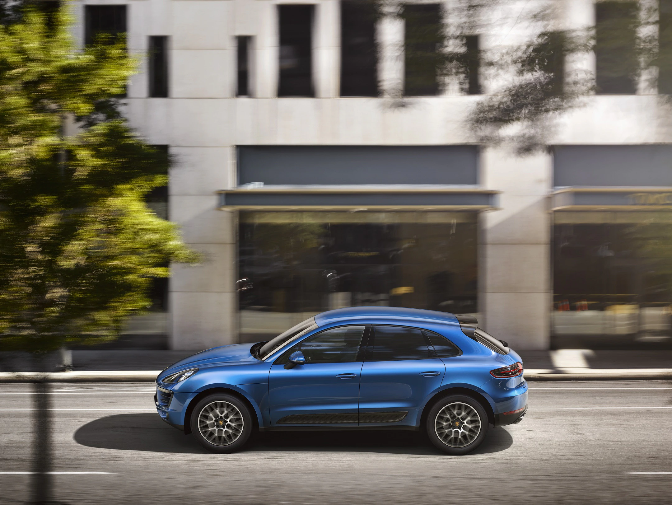 Blauer SUV fährt auf städtischer Straße, unscharfer Baum im Vordergrund, moderne Gebäude im Hintergrund.