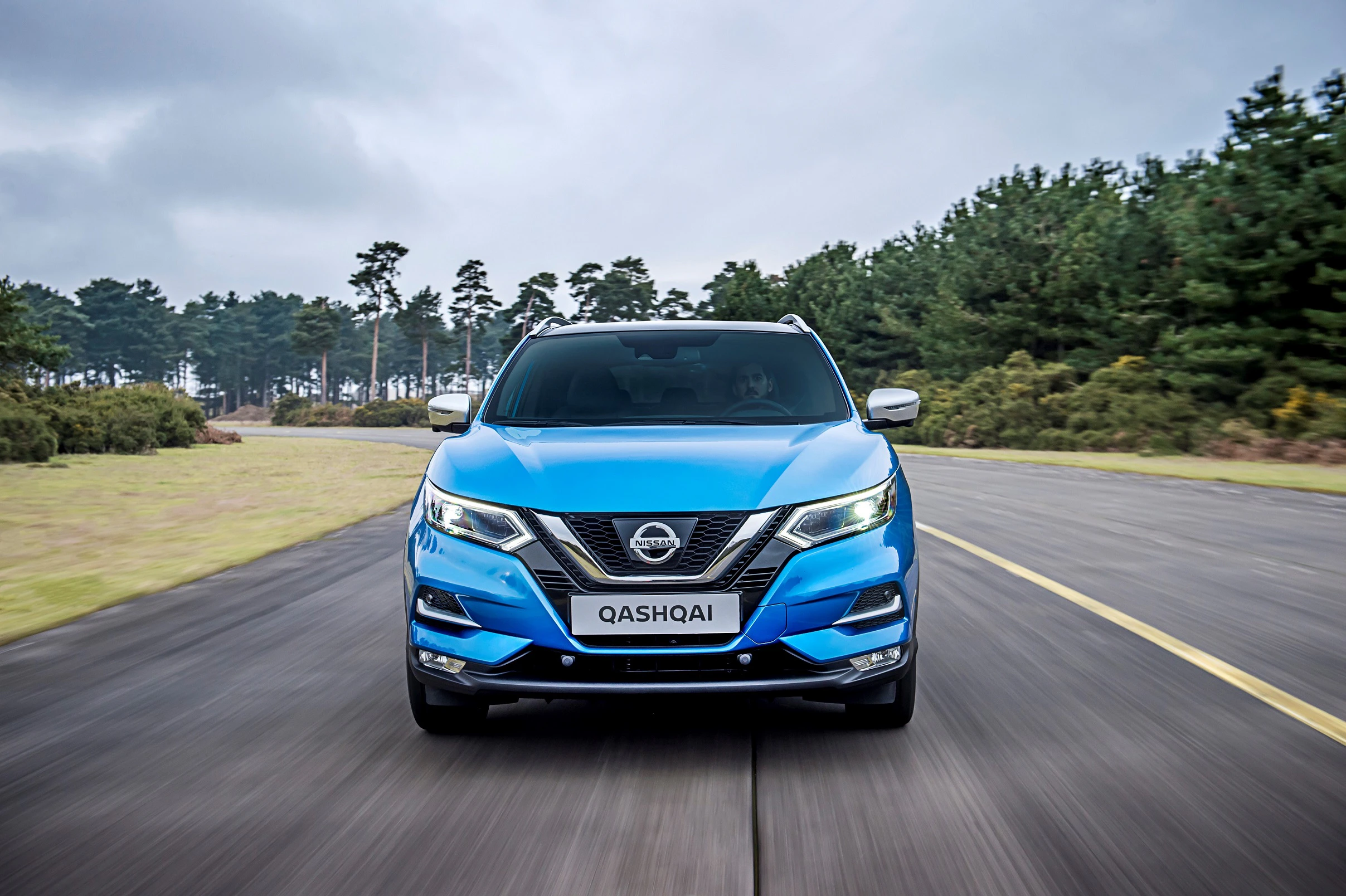 Blauer Nissan Qashqai fährt auf einer Landstraße, umgeben von Bäumen unter bewölktem Himmel. Frontalansicht des Fahrzeugs.