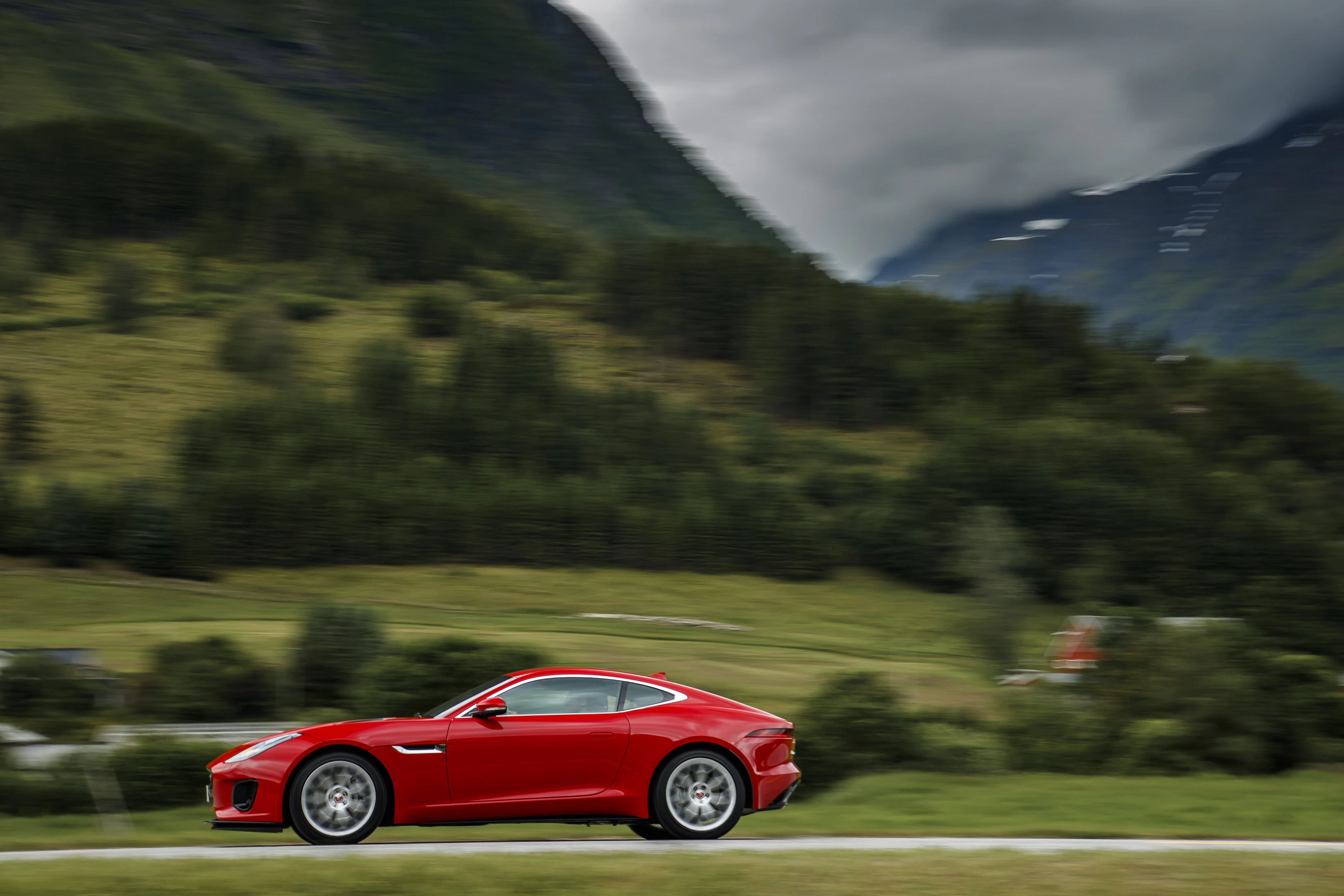 Ein rotes Sportauto fährt schnell über eine Landstraße, umgeben von grünen Hügeln und bewaldeten Bergen unter bewölktem Himmel.