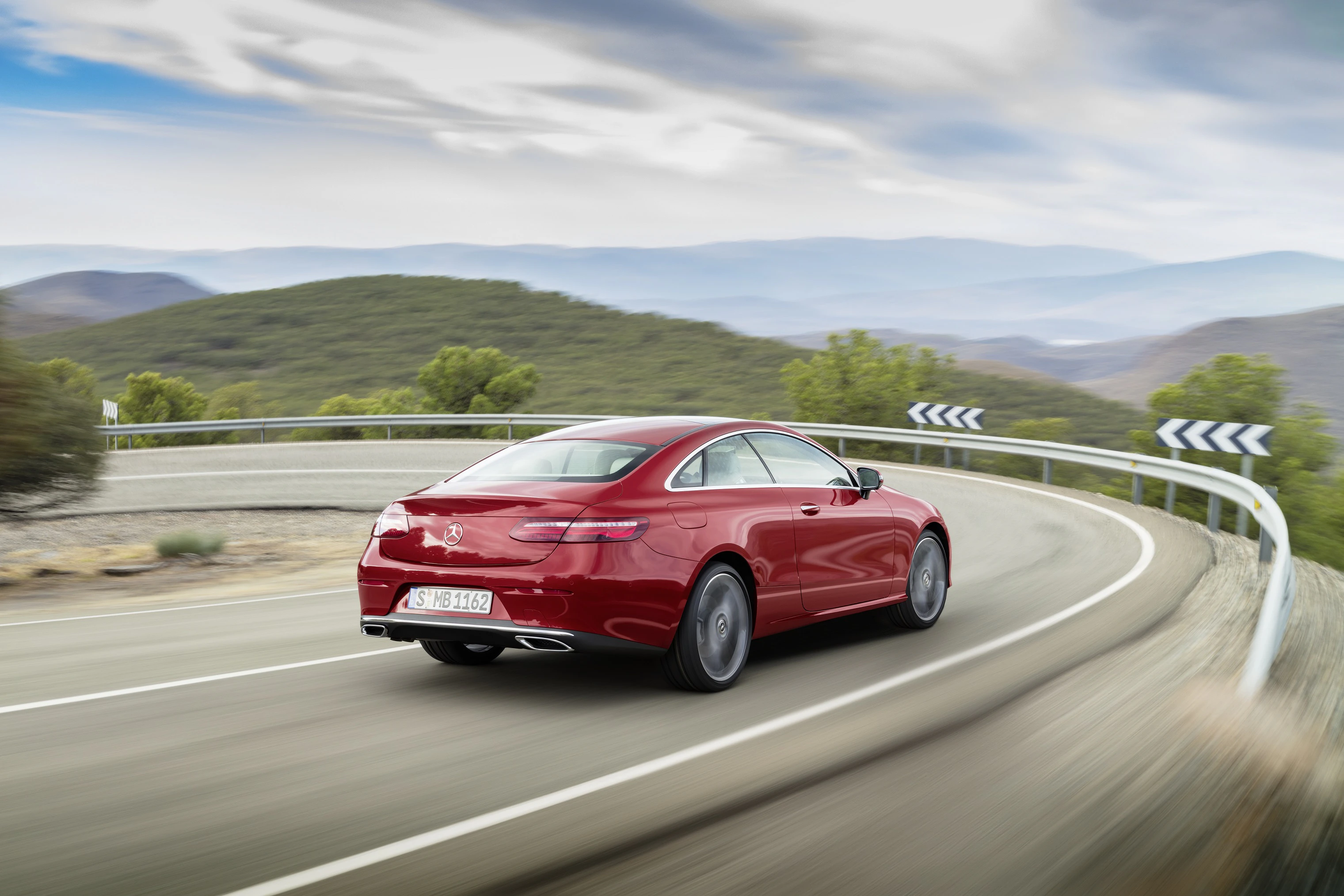 Roter Sportwagen fährt dynamisch auf kurviger Landstraße durch hügelige Landschaft, Himmel leicht bewölkt.
