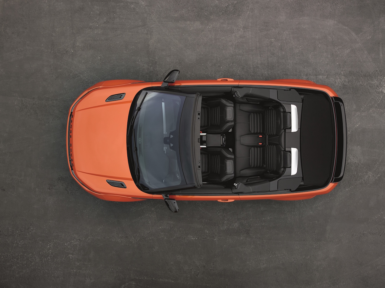 Orangefarbenes Cabrio von oben, mit geöffnetem Verdeck, auf grauem Untergrund. Vier schwarze Sitze sichtbar.