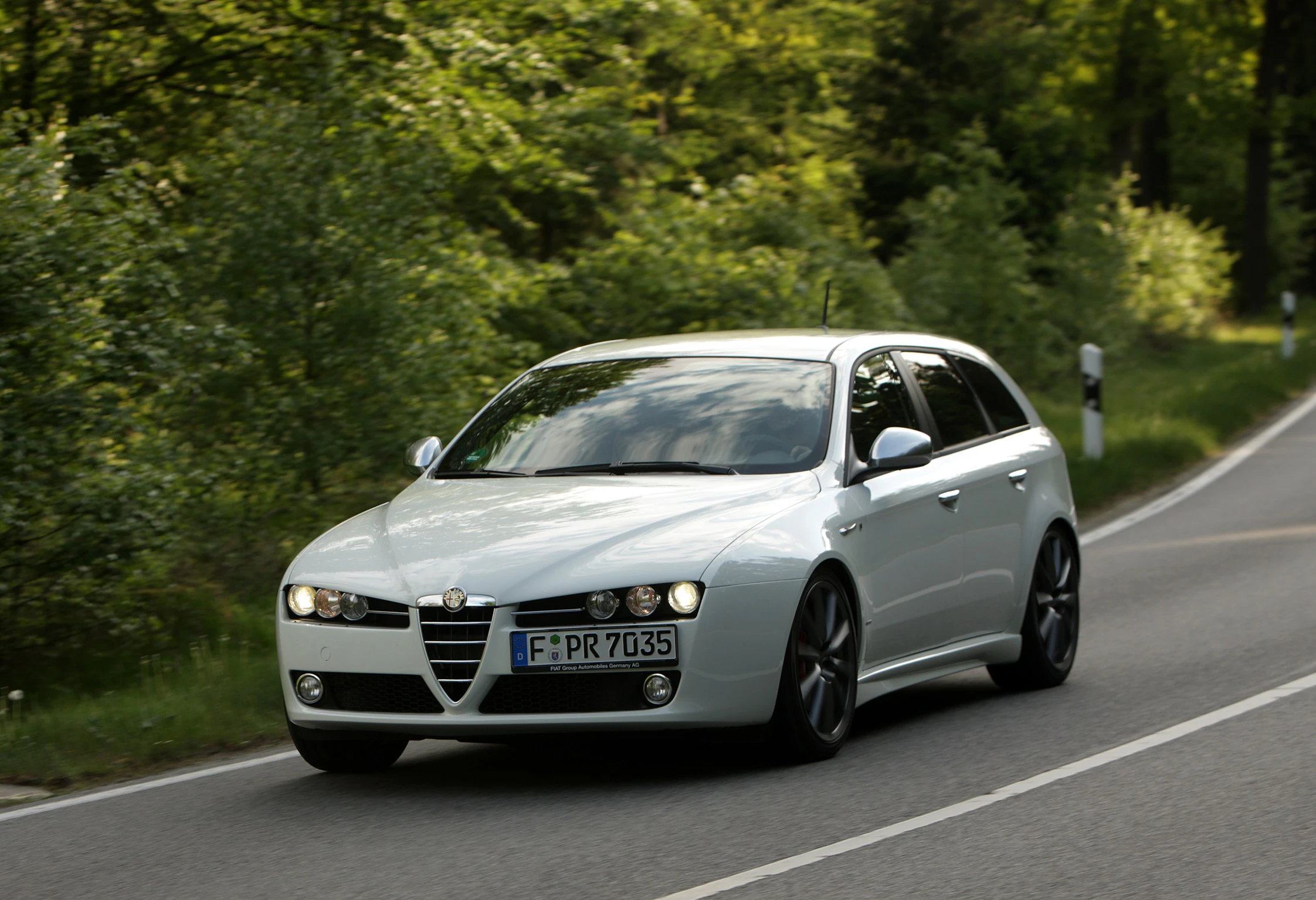 Silberner Alfa Romeo fährt auf kurviger Landstraße durch bewaldete Landschaft. Sonnenschein und grünes Laub umgeben die Strecke.