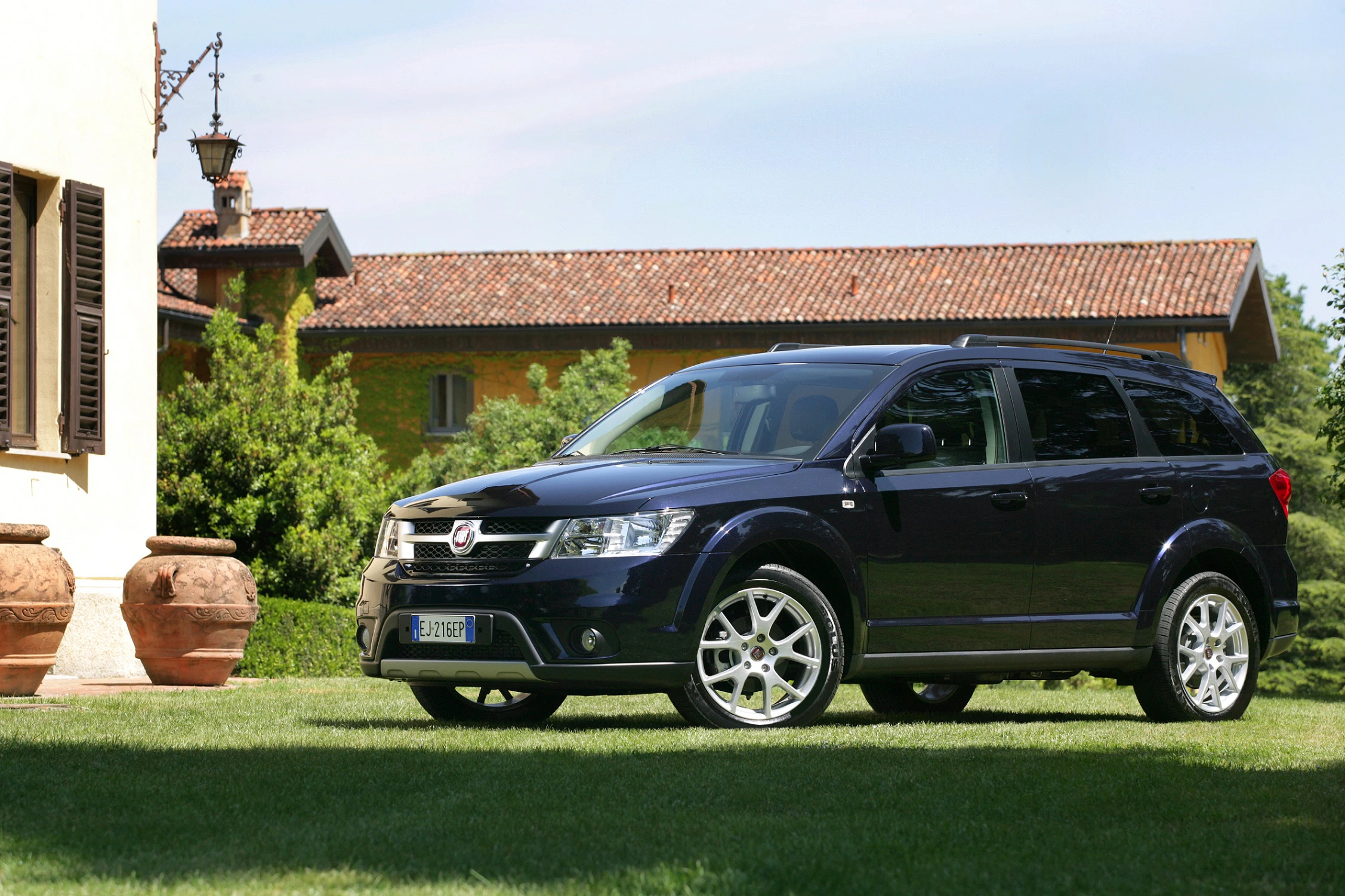Schwarzer SUV parkt auf Rasen vor einem gelben Haus mit Terrakotta-Dach und Garten im Hintergrund.