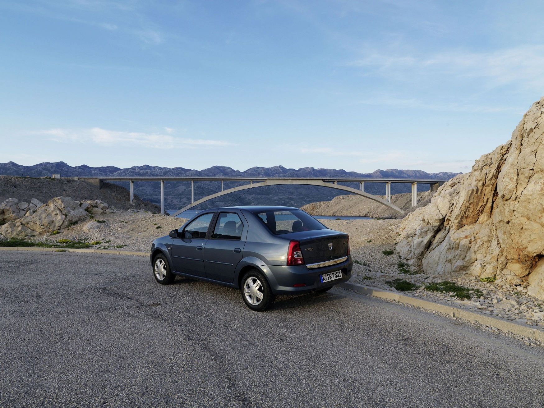 Ein graues Auto steht auf einer Straße vor einer felsigen Landschaft und einer Brücke, im Hintergrund sind Berge unter klarem Himmel zu sehen.