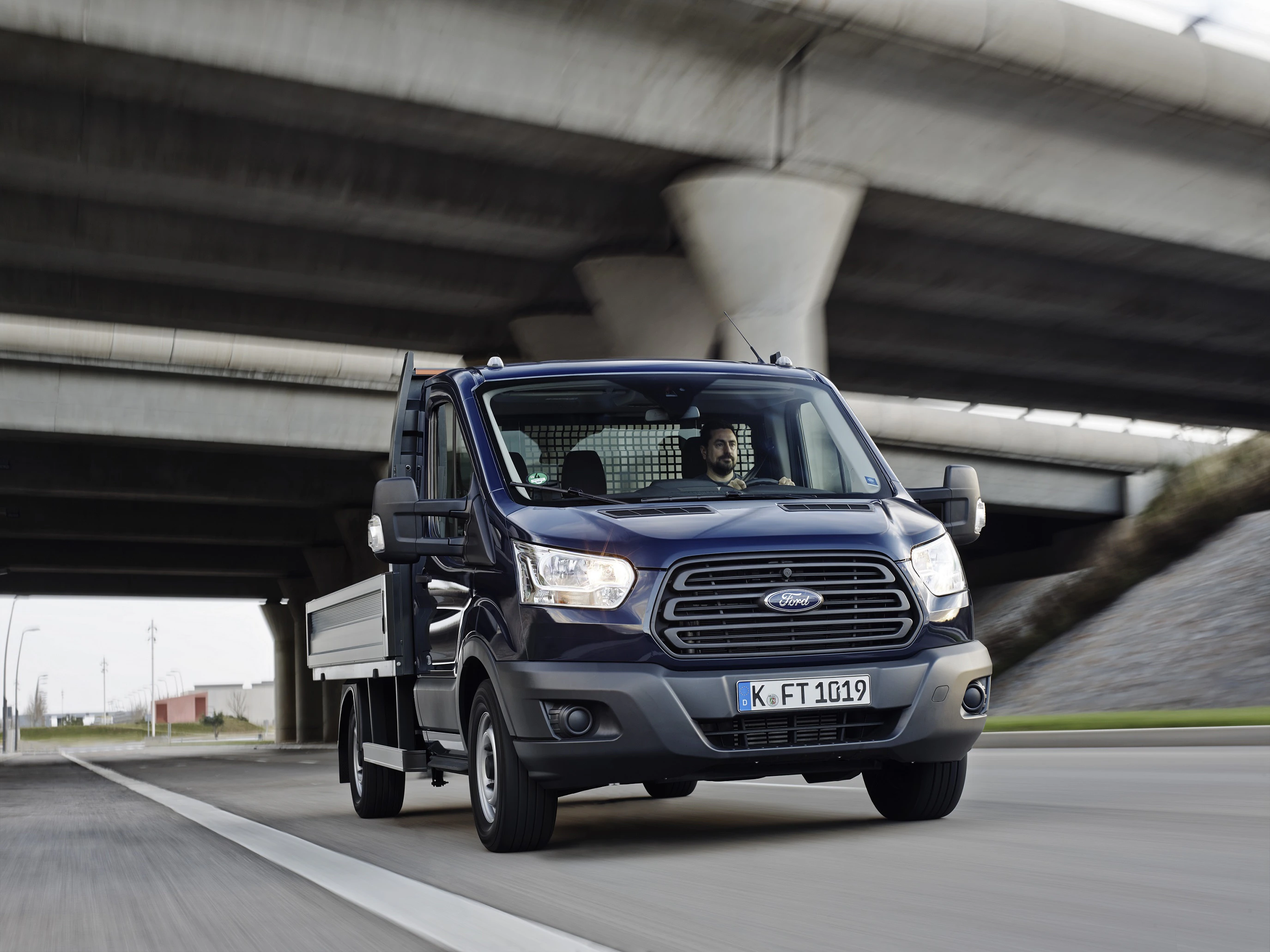 Ein dunkelblauer Ford-Transporter fährt auf einer Straße unter einer Betonbrücke hindurch. Der Fahrer blickt konzentriert nach vorn.