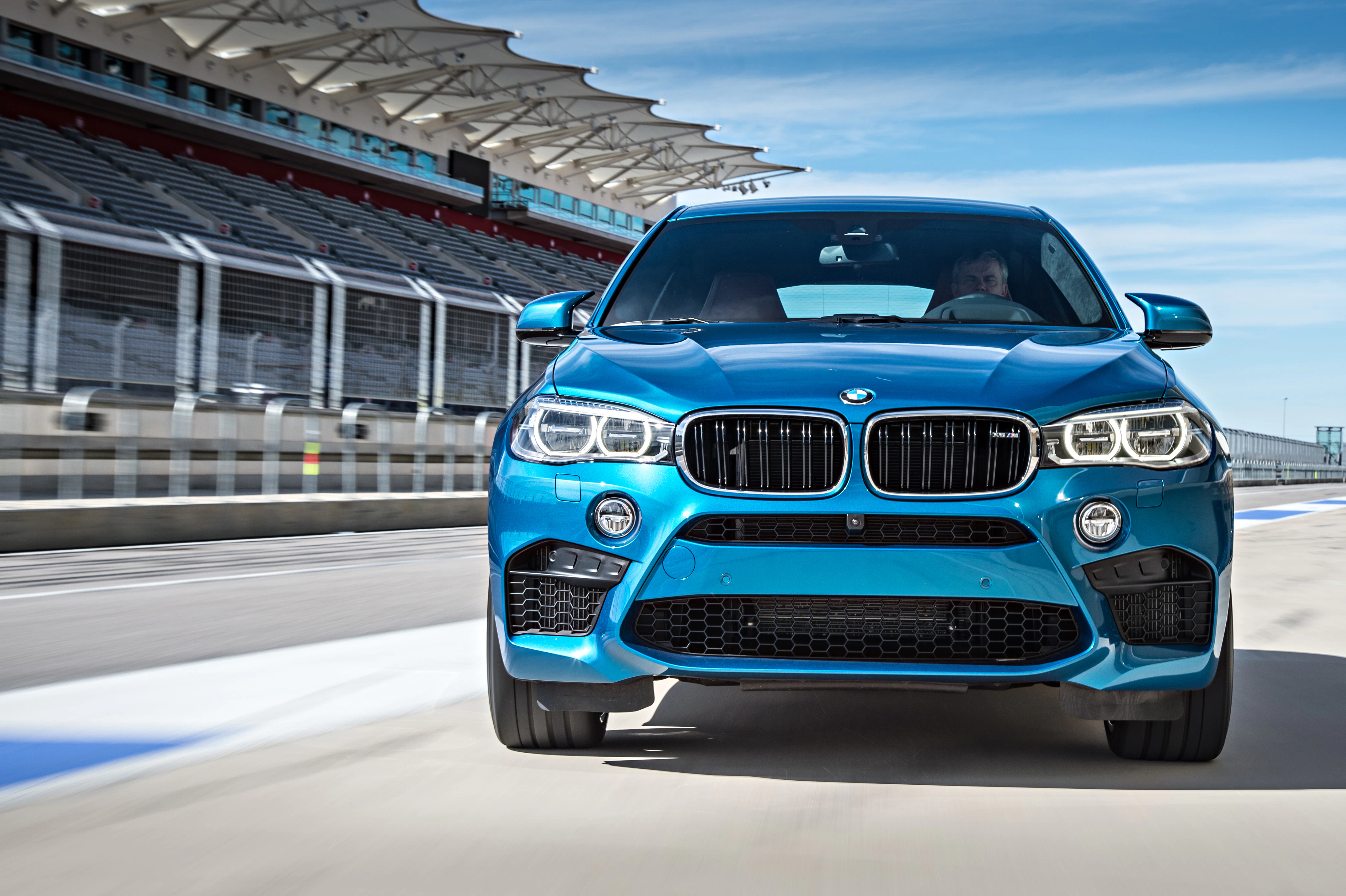 Ein blauer BMW SUV fährt auf einer Rennstrecke mit leeren Tribünen im Hintergrund bei sonnigem Wetter.