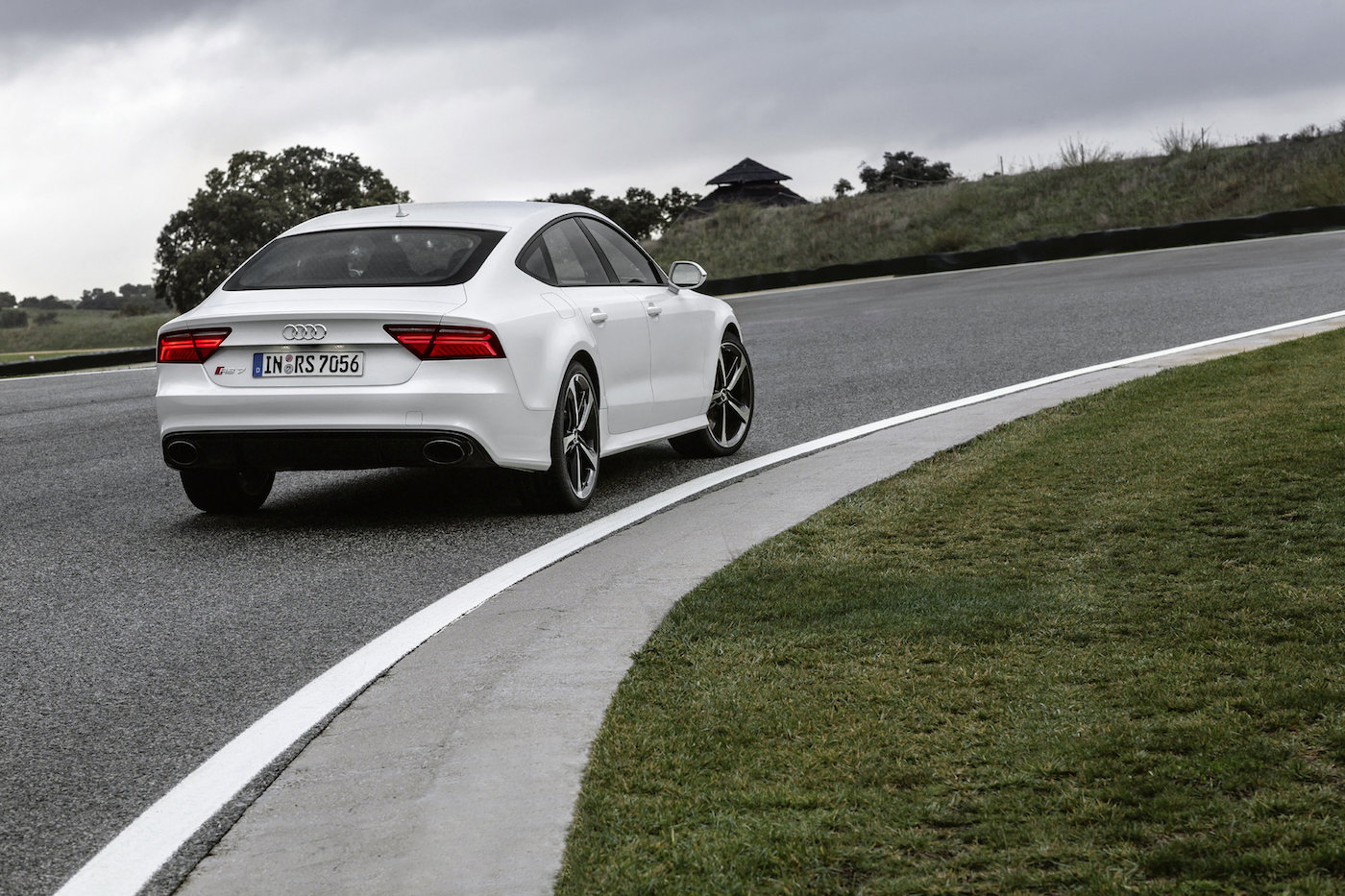Weißer Audi RS7 fährt auf einer kurvigen Landstraße, umgeben von grüner Landschaft und bewölktem Himmel.