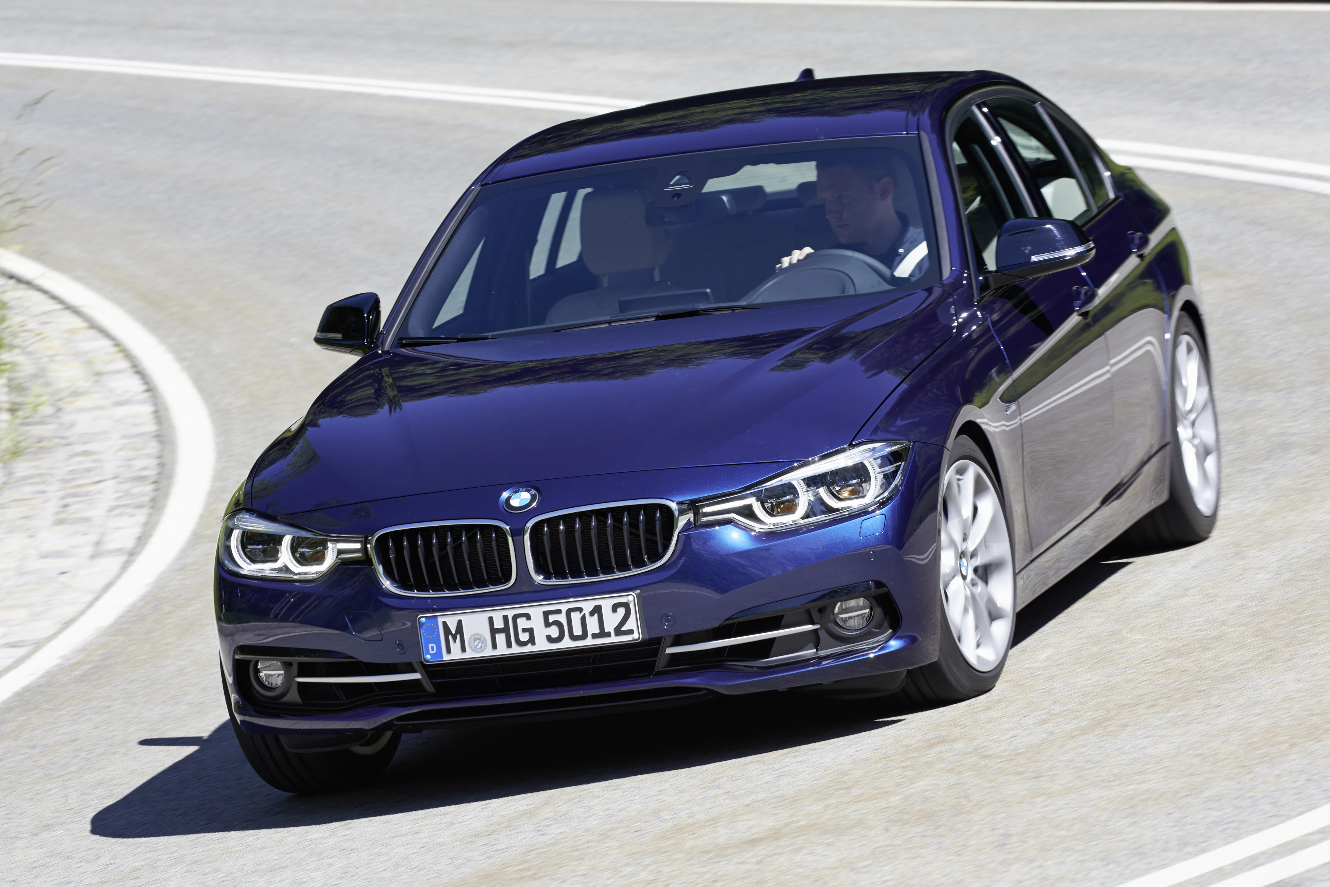 Blauer BMW fährt dynamisch eine kurvige Straße entlang, Fahrer im Inneren sichtbar. Lizenznummer: M-HG 5012.