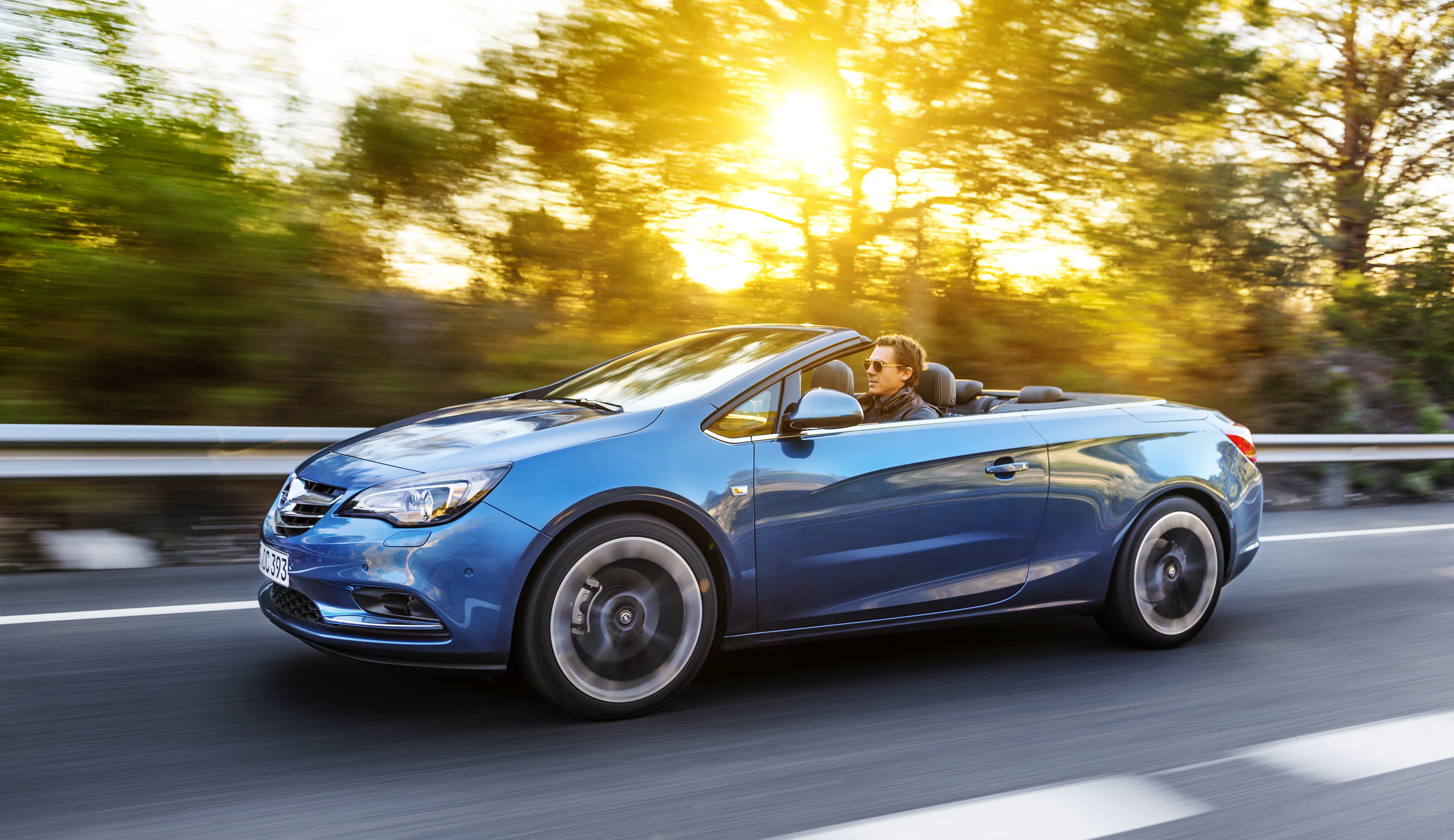 Blaues Cabrio fährt bei Sonnenuntergang auf einer Landstraße, umgeben von verschwommenen Bäumen; Fahrer mit Sonnenbrille entspannt.