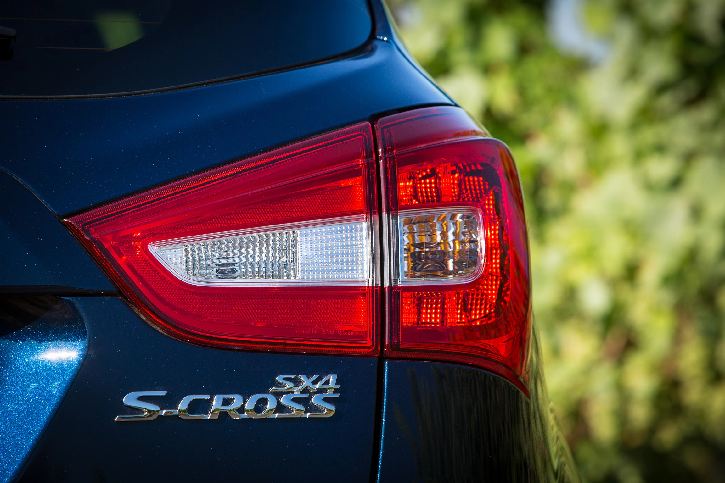 Nahaufnahme eines blauen Suzuki SX4 S-Cross mit Fokus auf das Rücklicht und das Modellabzeichen, vor unscharfem, grünem Hintergrund.