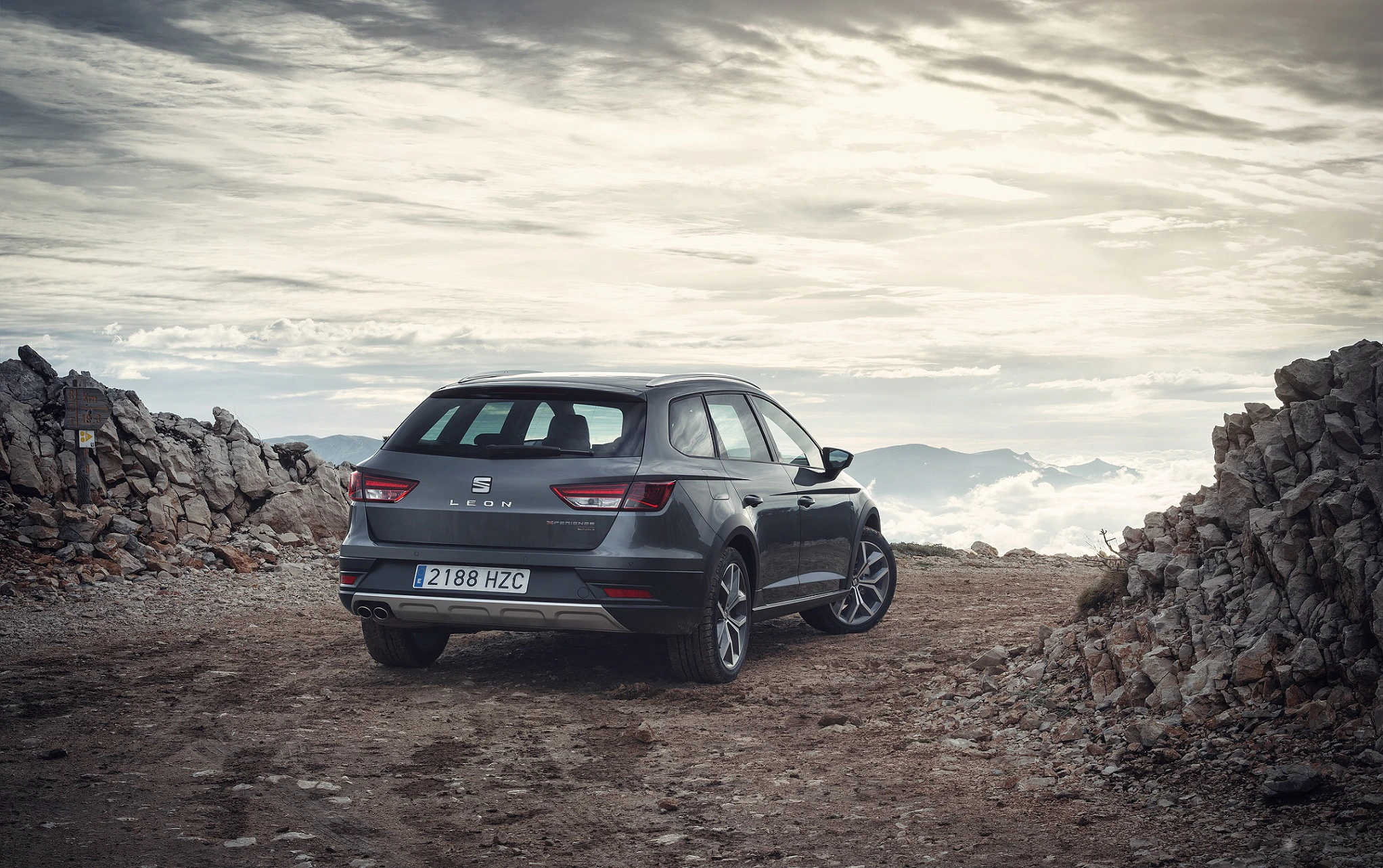 Ein dunkler SUV steht auf einem felsigen Bergweg mit wolkigem Himmel und Bergpanorama im Hintergrund.