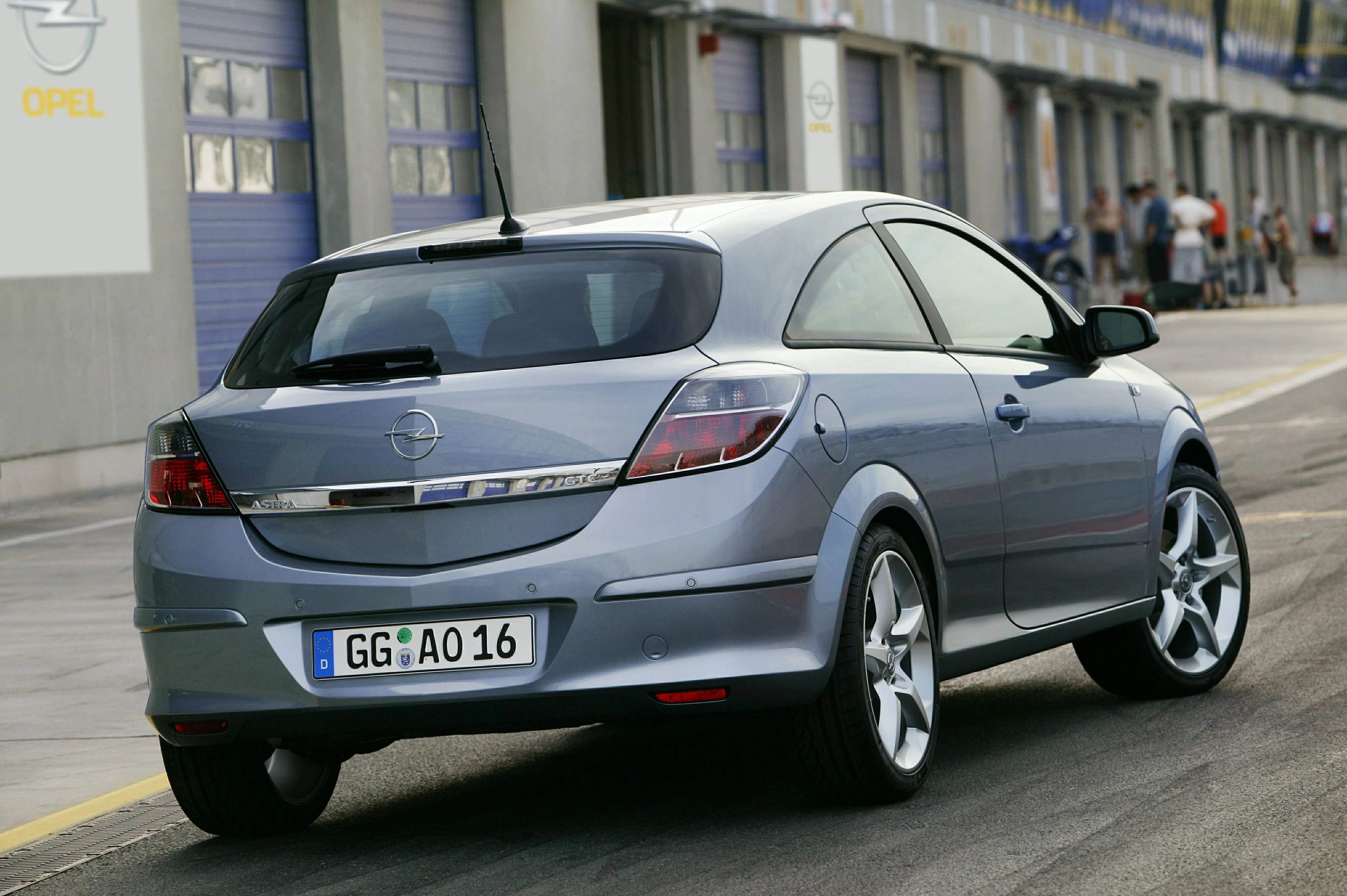 Silberner Opel Astra GTC in einem Parkbereich vor einer Werkstatt oder Garage, von hinten und leicht seitlich aufgenommen.